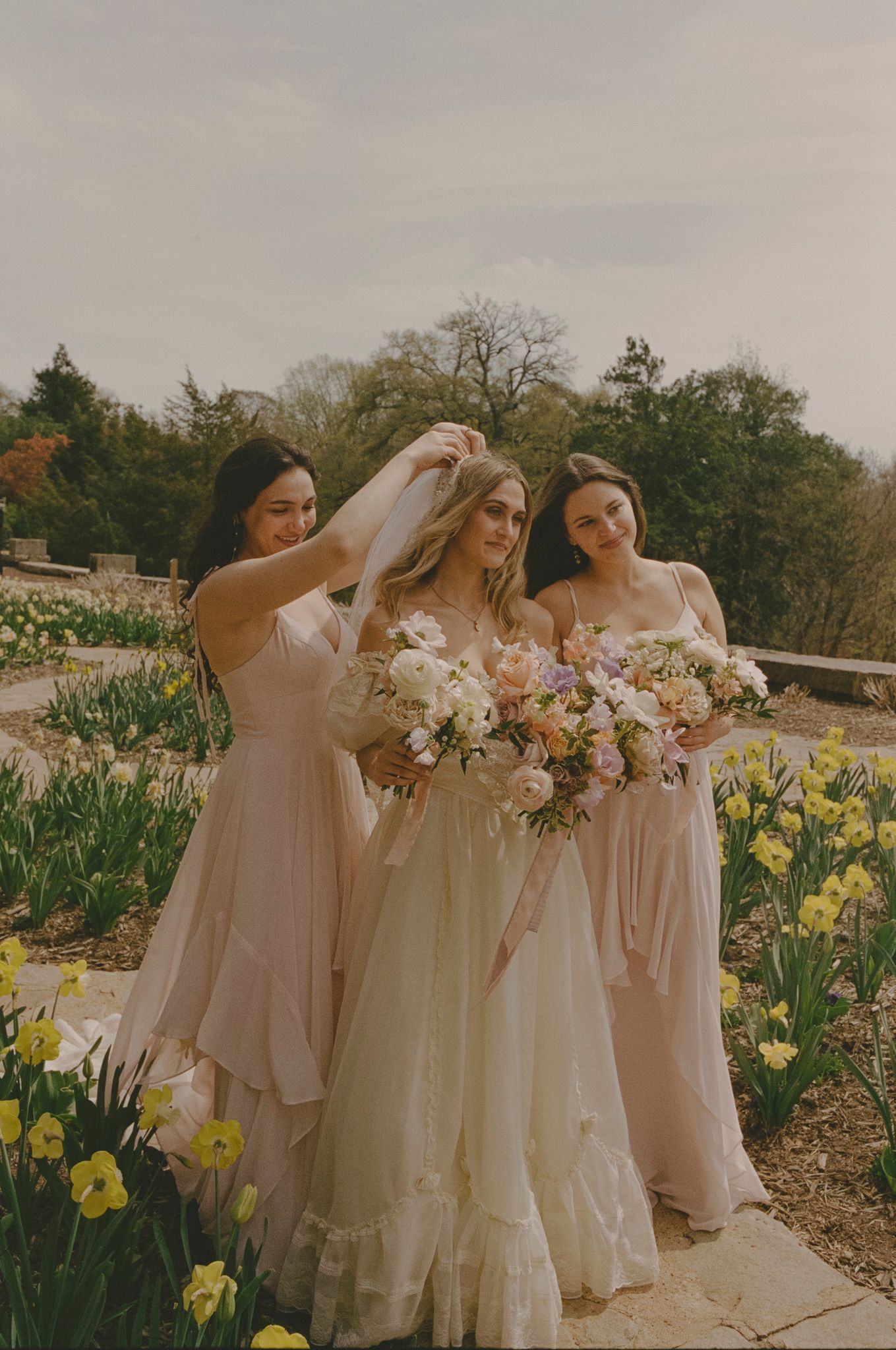 Bridesmaids in blush pink dresses pose together in a garden filled with blooming yellow daffodils.