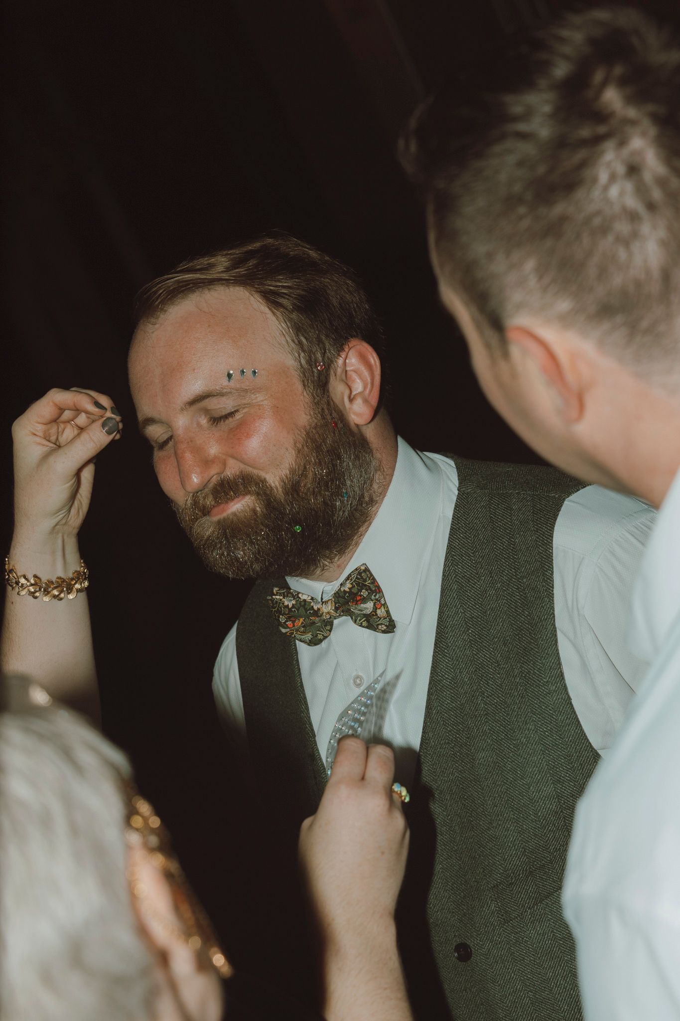 A bearded guest at a formal event wearing a bow tie and vest has his boutonniere adjusted in a dimly lit setting.