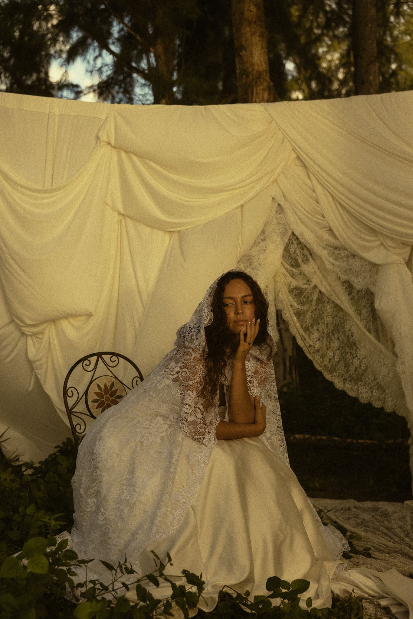 Bride in white lace gown and veil sits pensively beneath draped fabric canopy outdoors among trees.