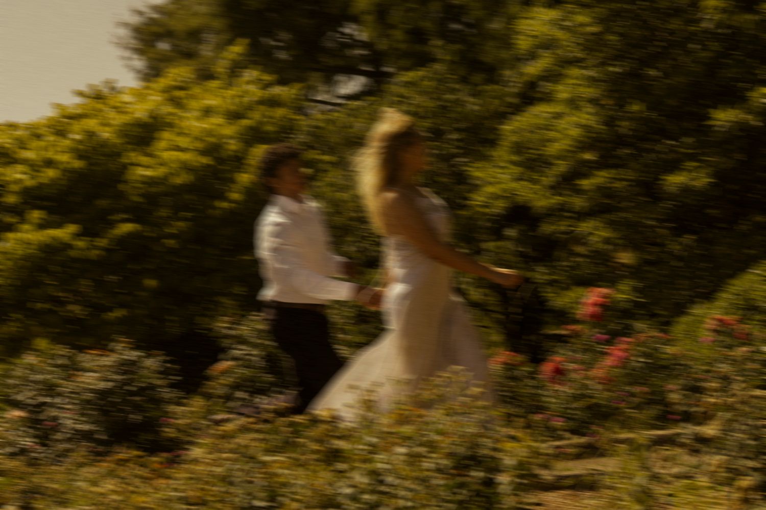 Blurred motion shot of bride and groom running through lush green garden on wedding day.