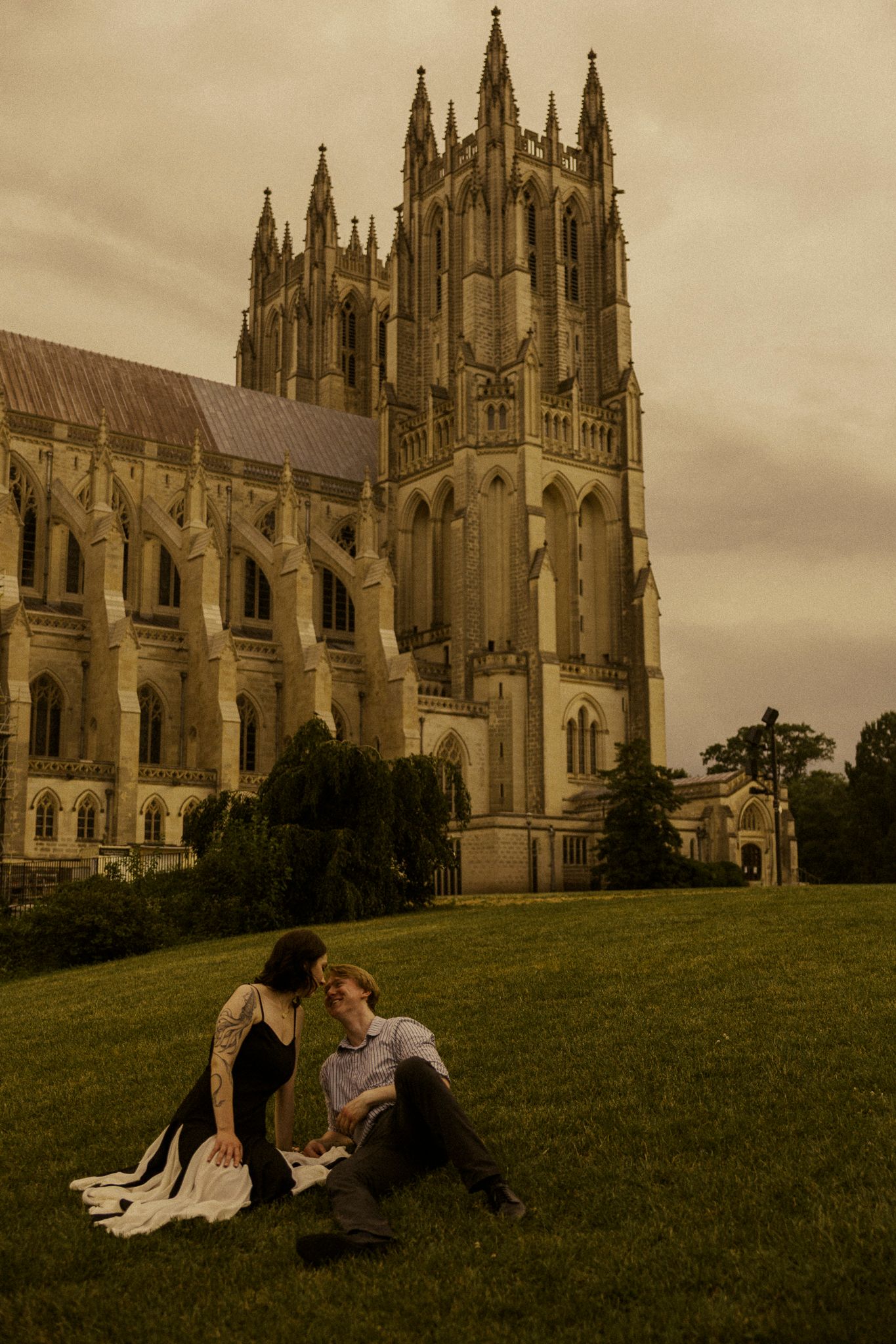 Couple sitting on grass before Washington National Cathedral's Gothic towers under dramatic cloudy skies.