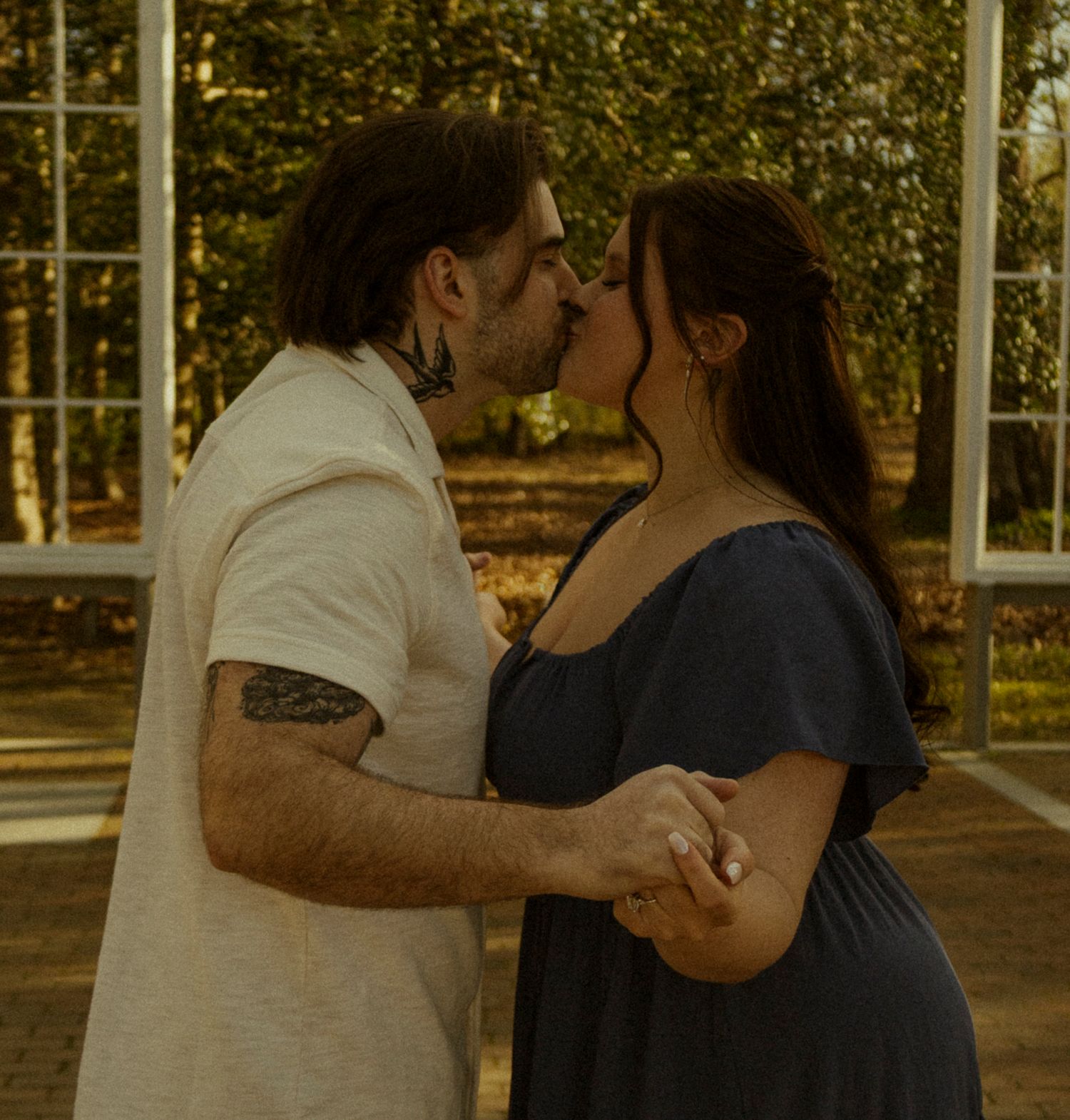 Romantic couple sharing a kiss outdoors near white window frames with lush green trees in the background.