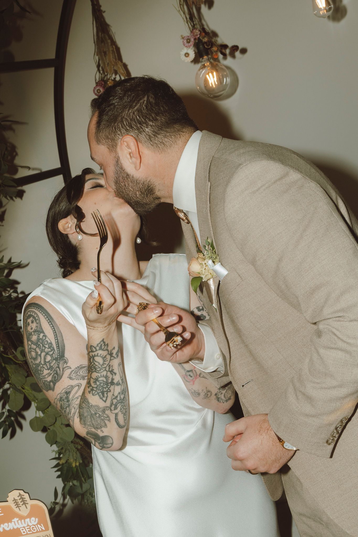 A couple shares a romantic kiss during their wedding celebration, with the bride wearing white and groom in beige.