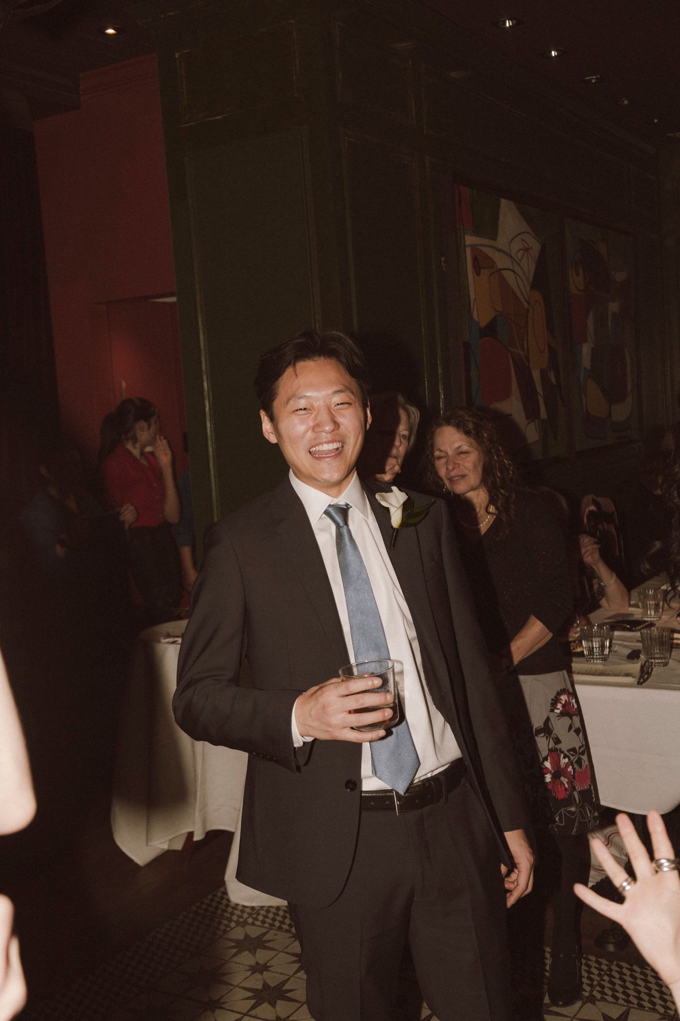 Smiling groom in dark suit and blue tie holding a drink at his wedding reception celebration.