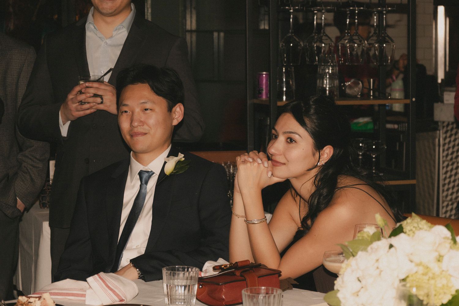Couple in formal attire seated at wedding reception table, listening attentively with white floral centerpiece nearby.