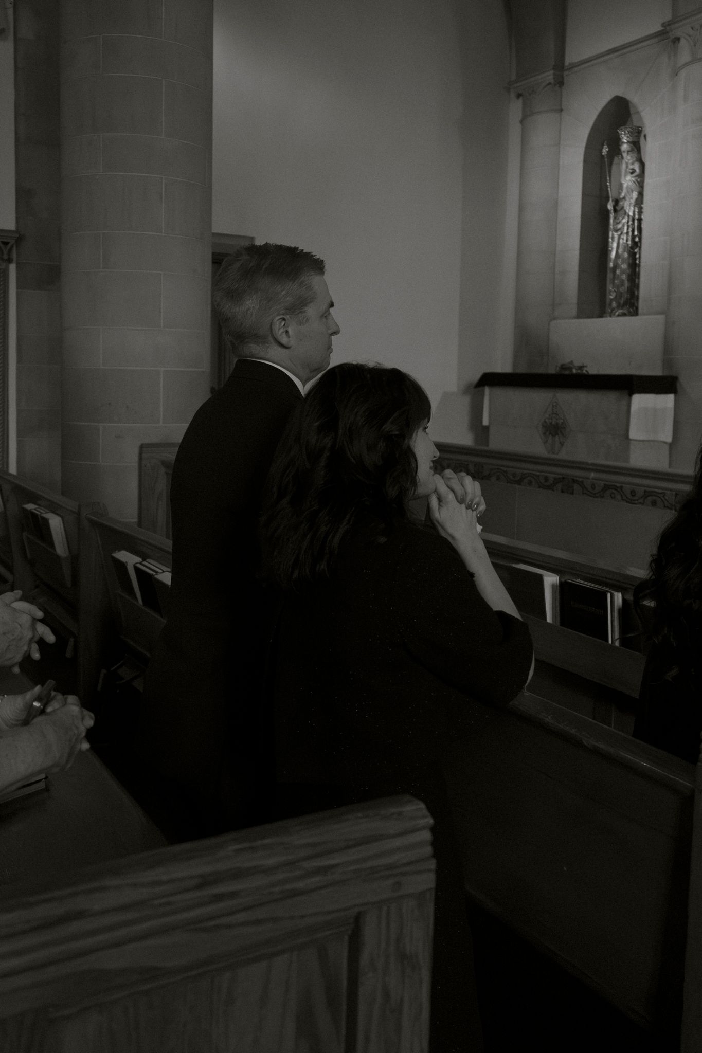 Black and white photo of man and woman kneeling in prayer at wooden church pew.