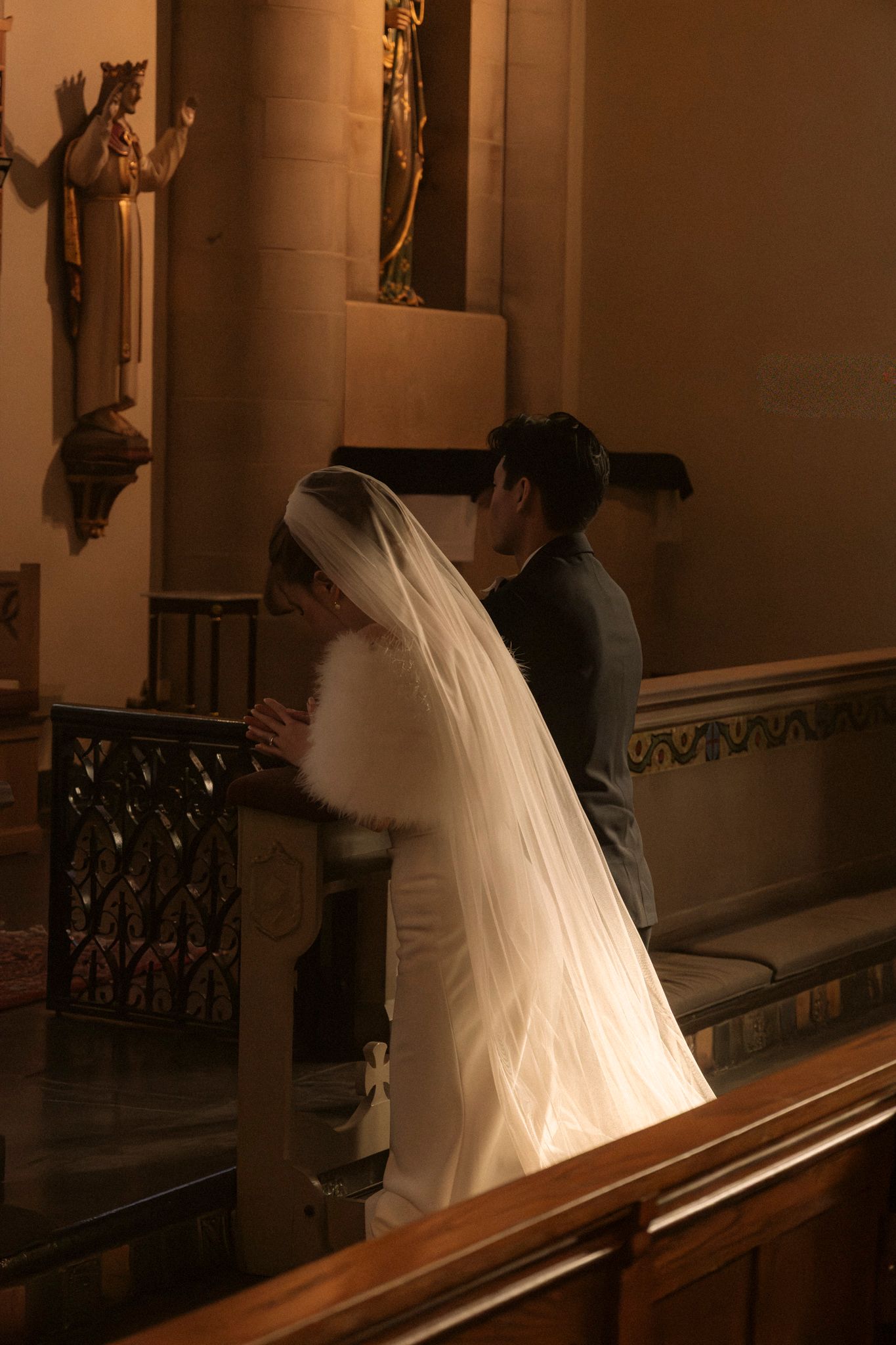 Bride in white wedding gown and veil praying at church altar rail.