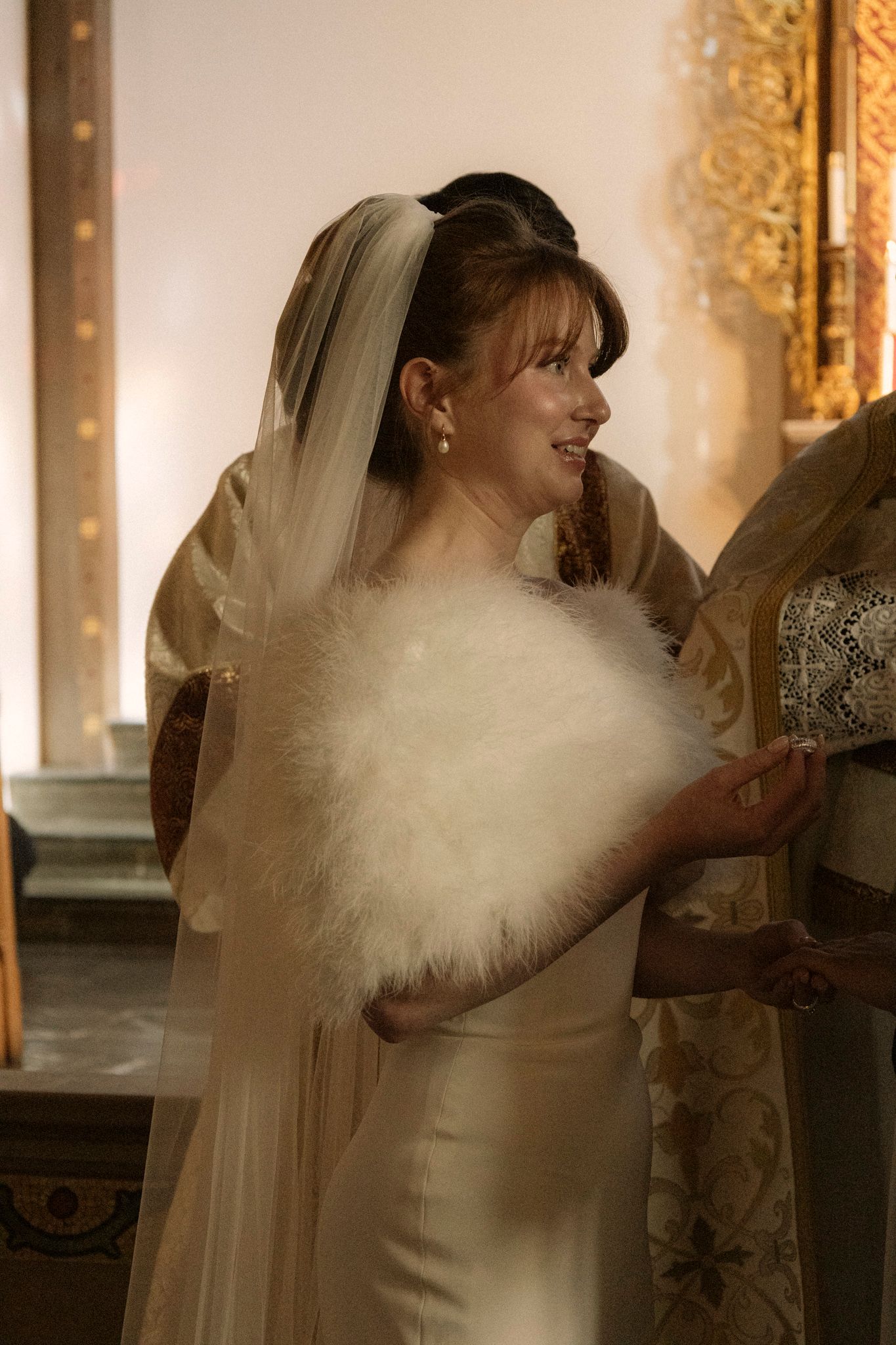 Smiling bride in satin gown with veil and white feather wrap stands near golden church altar.