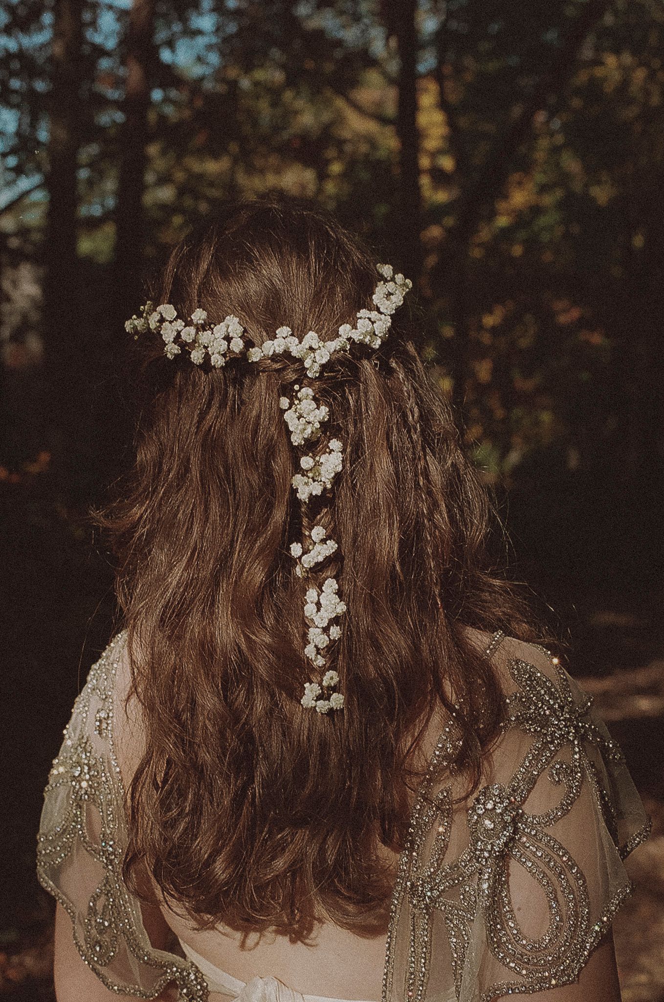 A person with wavy brown hair adorned with delicate white flowers poses against a dark forest background.