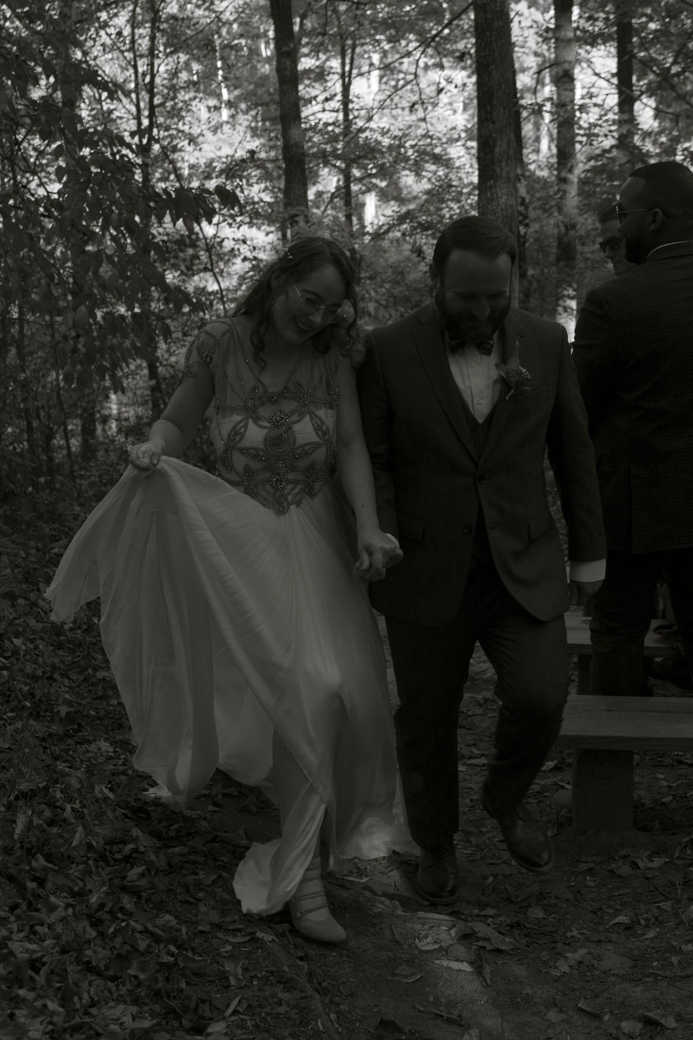 A couple walks together through a wooded forest path in black and white, with the bride holding up her flowing white dress.