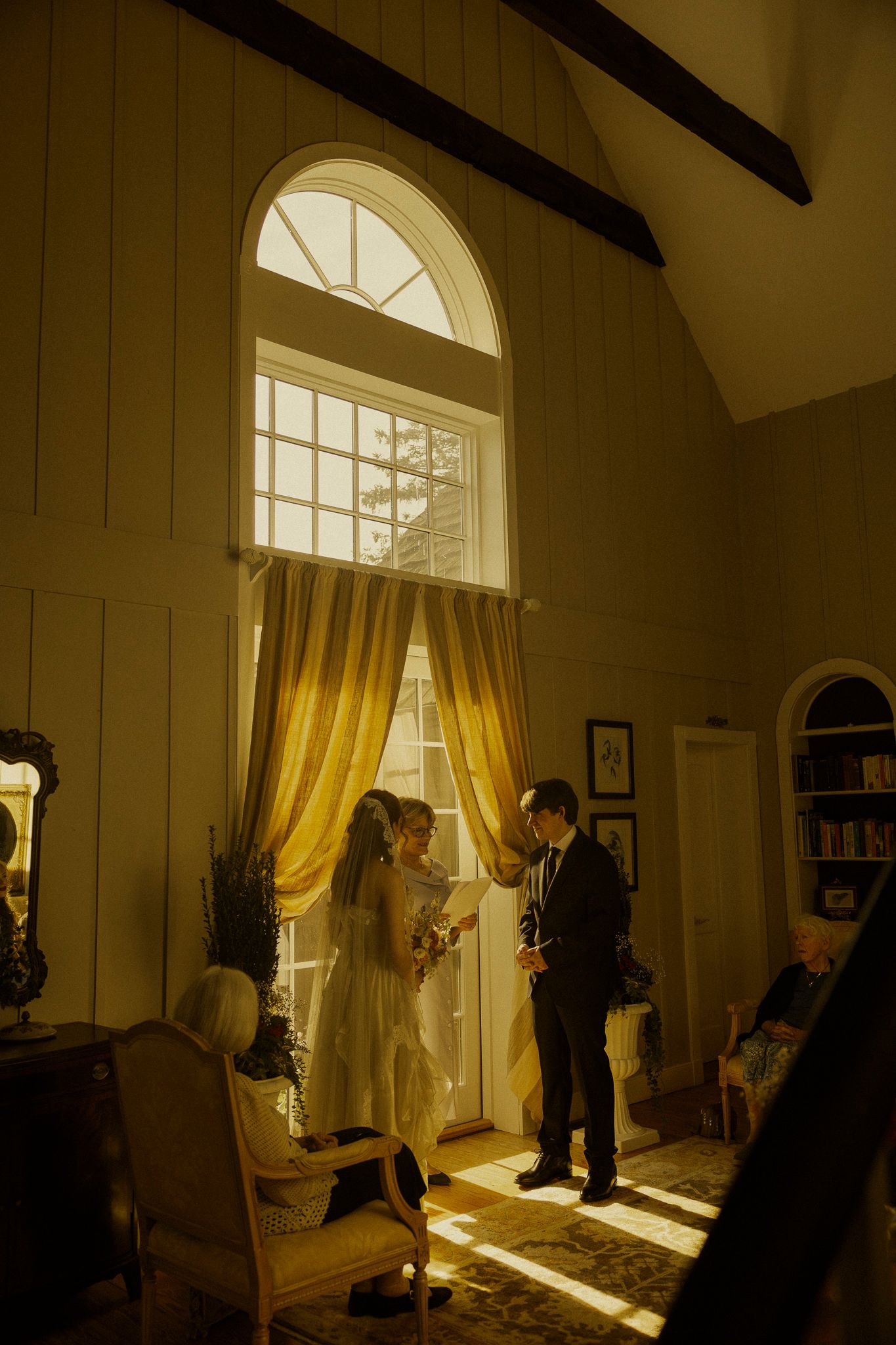 Bride and groom exchange vows by sunlit arched windows with golden curtains in an elegant rustic chapel interior.