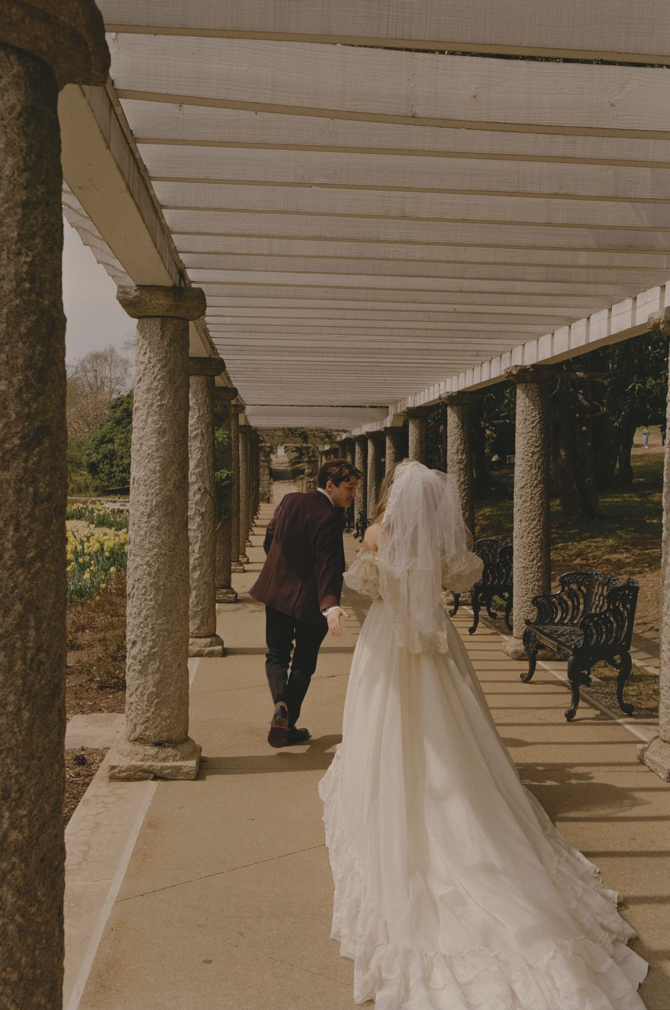 A person in a flowing white wedding dress walks along a covered outdoor colonnade with ornate stone pillars.