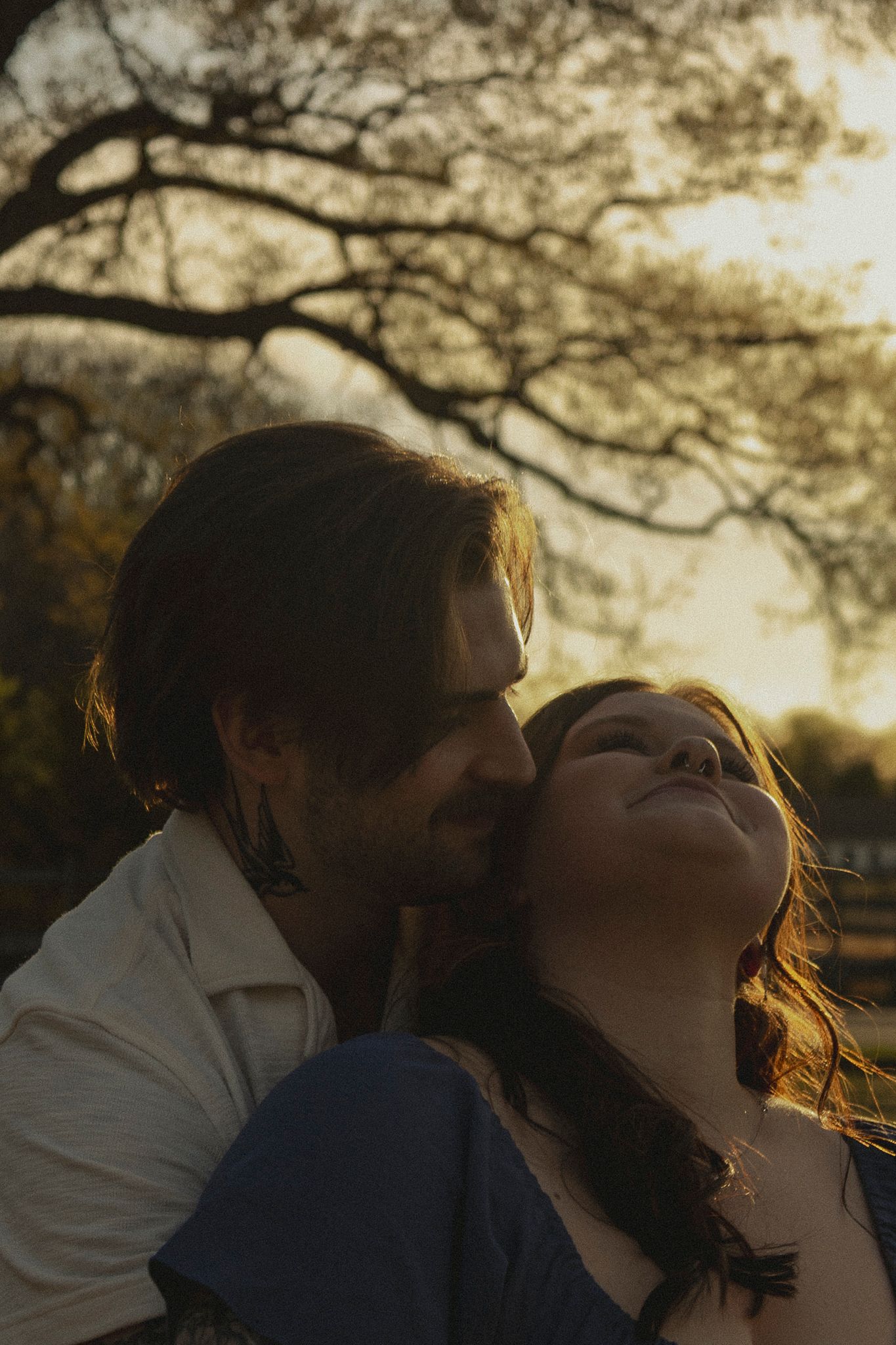A couple shares a tender romantic moment at golden hour, with the woman tilting her head back and smiling beneath a tree.