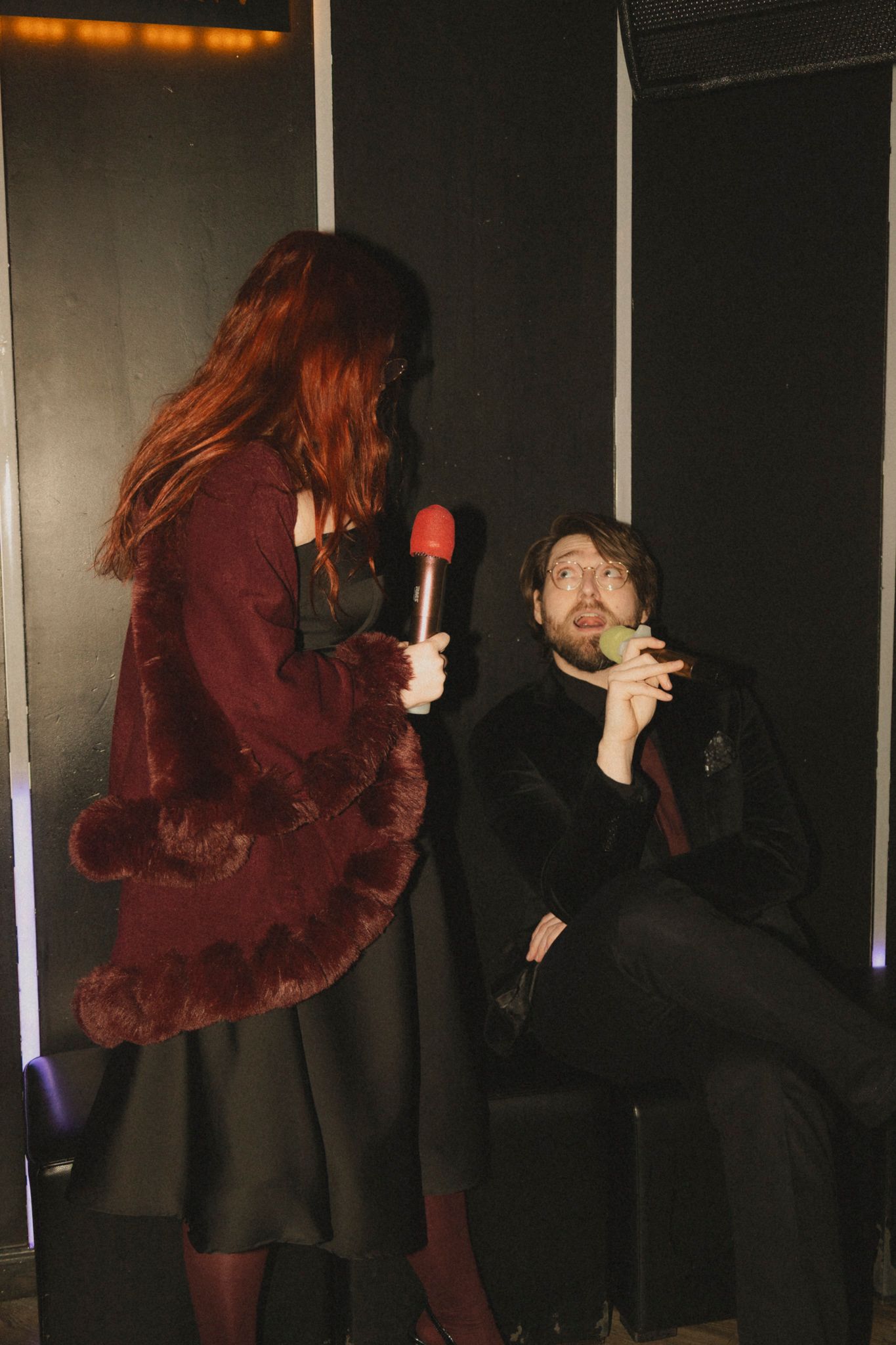 Two people in dark clothing interact playfully with a red lollipop in a dimly lit setting.