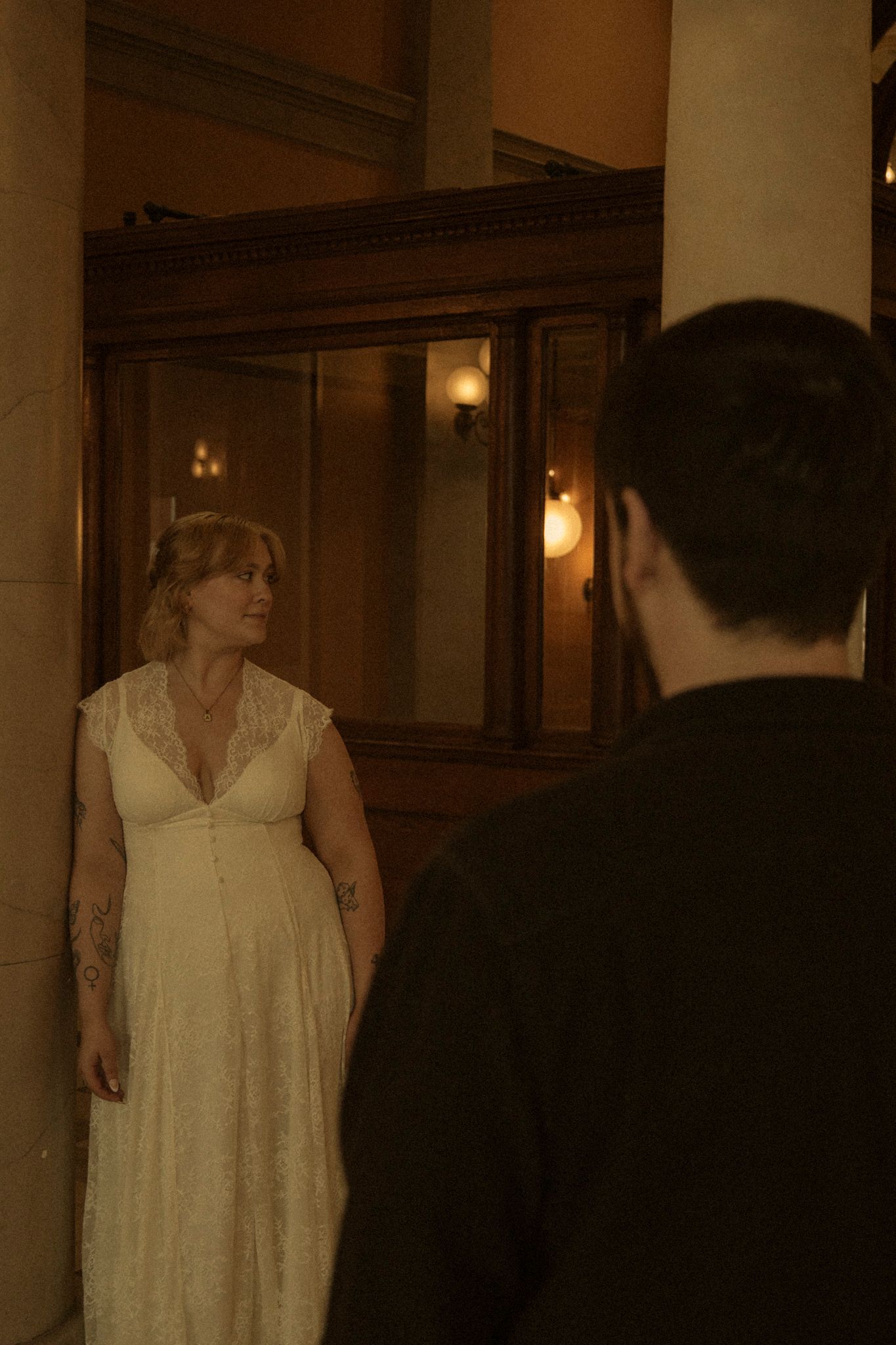 A couple in formal attire stand in a dimly lit hallway with vintage wall sconces casting a warm glow.