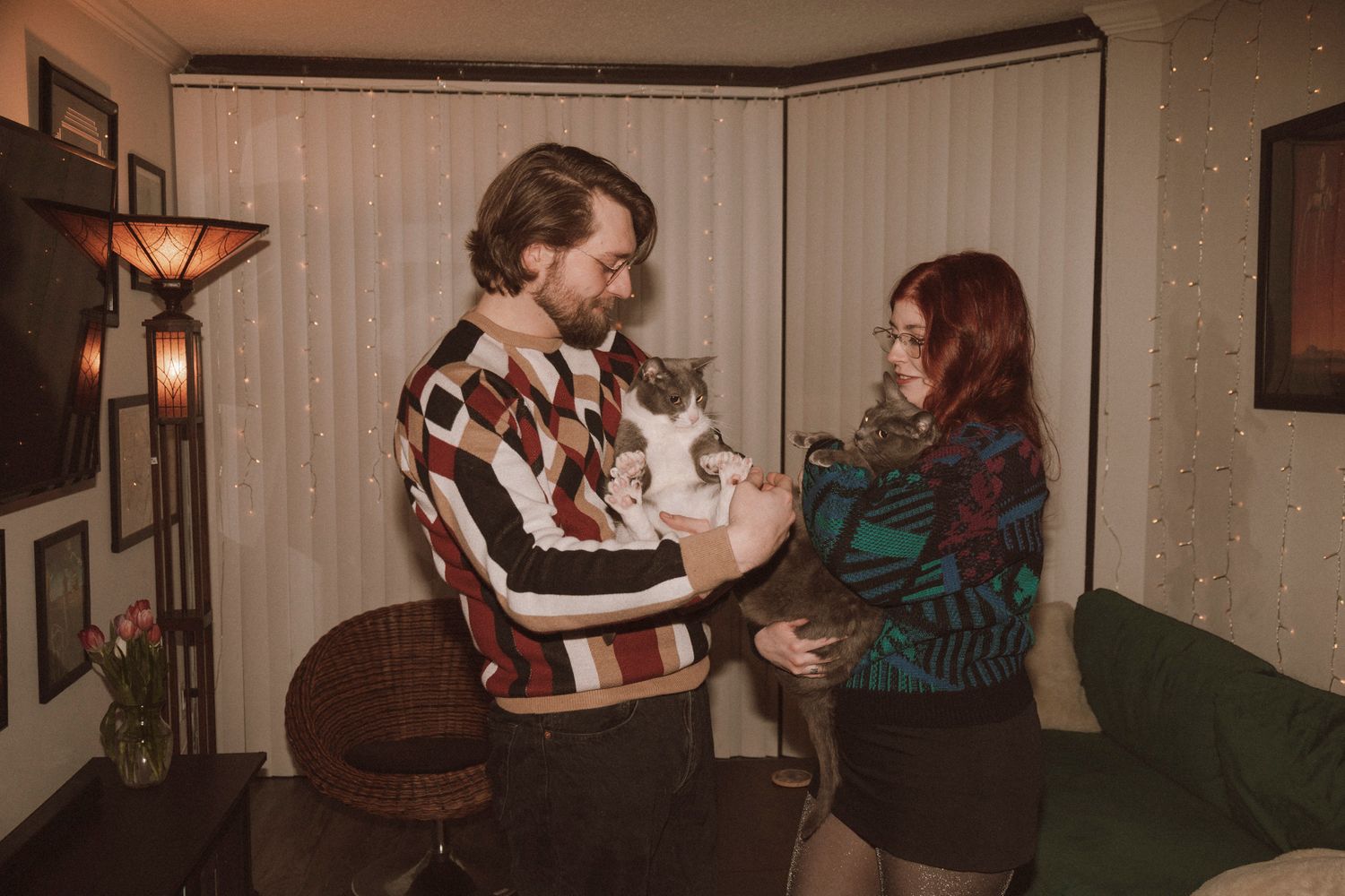 A couple sits on a green couch in a vintage-style living room while holding a gray and white cat.