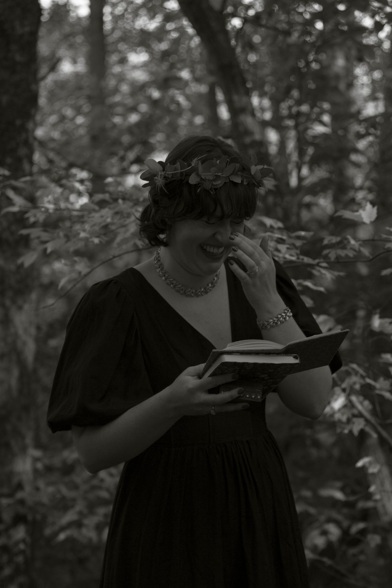 A person in a black dress reads a book while standing in a wooded area, shown in black and white photography.
