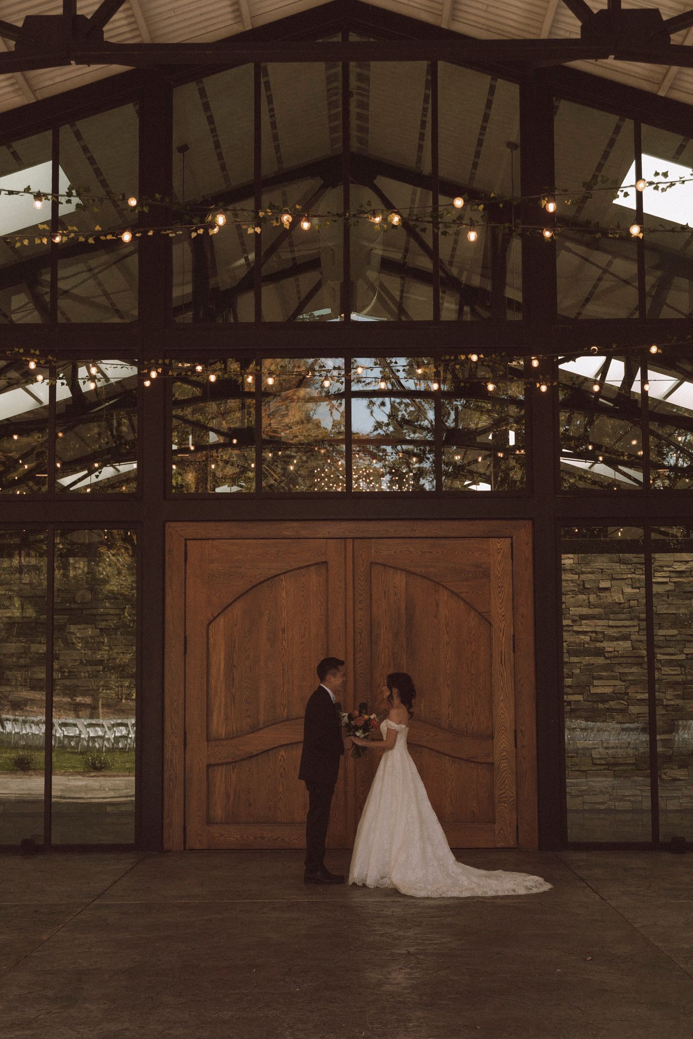 A couple stands silhouetted in front of large wooden doors under a rustic glass-roofed venue with string lights overhead.