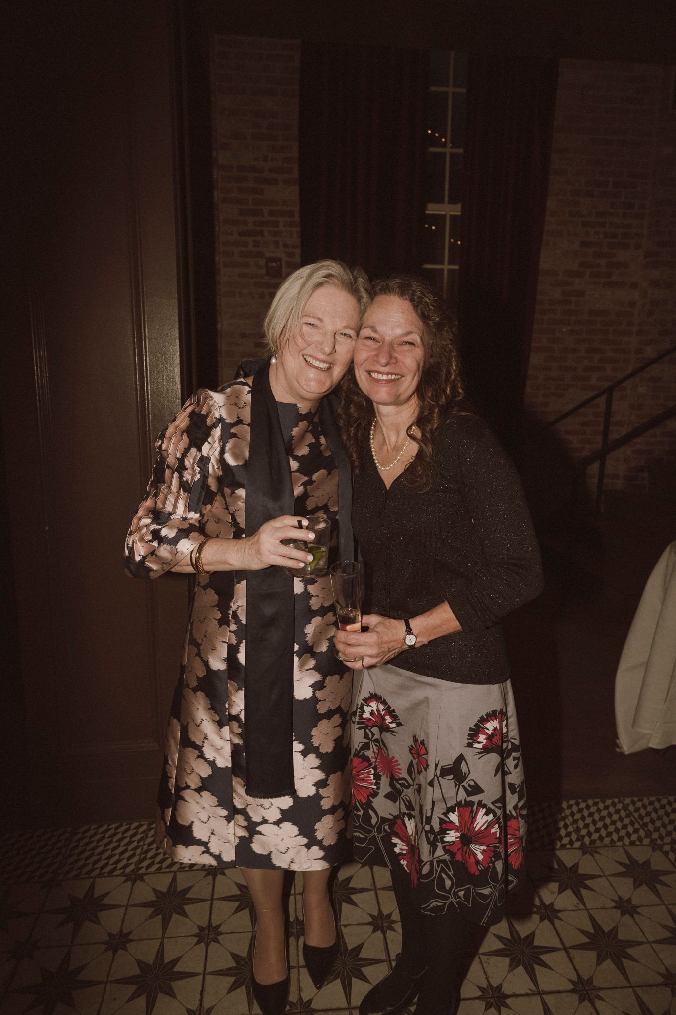Two women smiling and embracing at wedding reception, one in floral dress, one in floral skirt.