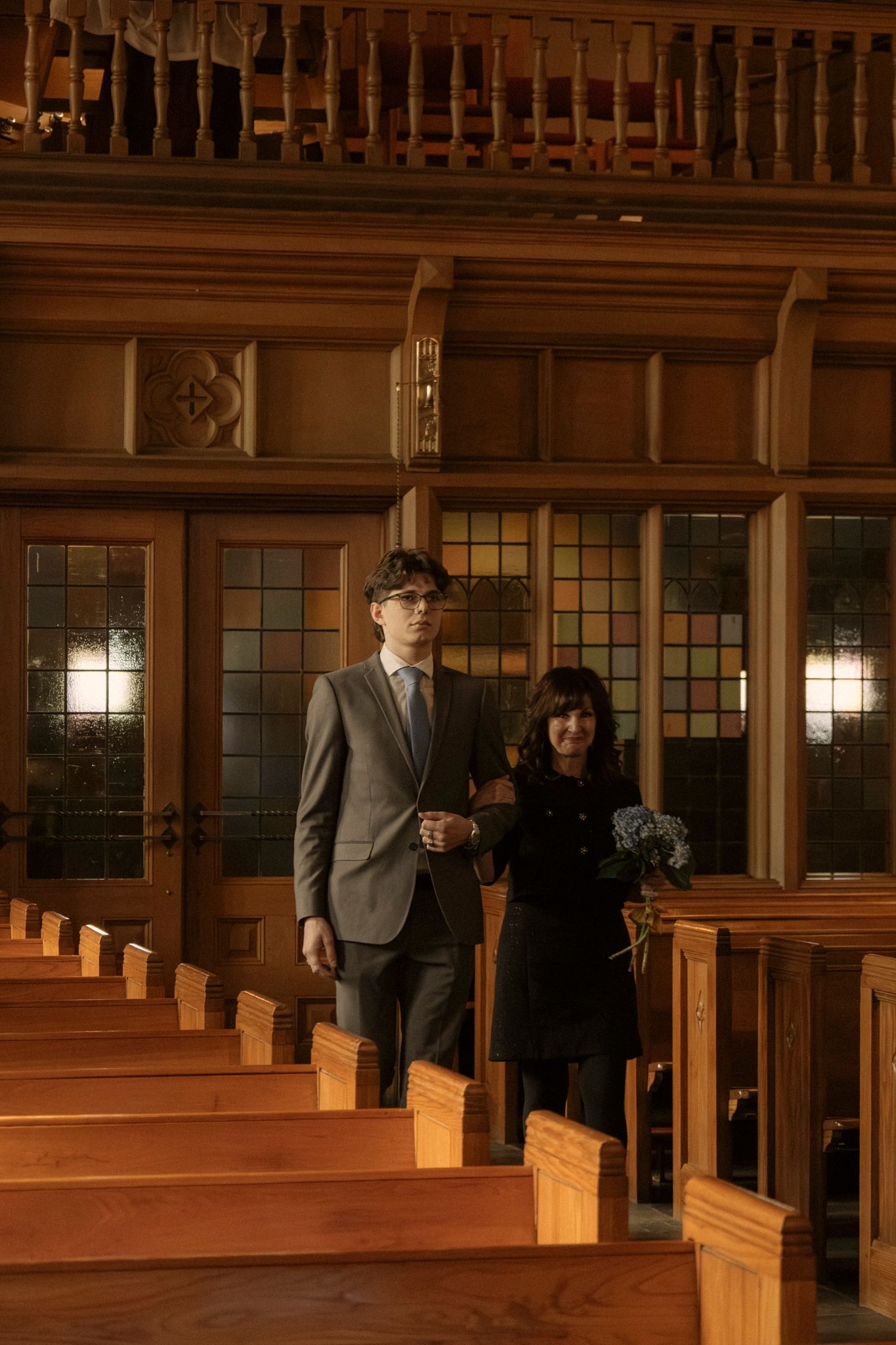 A couple walks down a church aisle, man in gray suit, woman in black dress holding flowers, amid warm wooden pews.