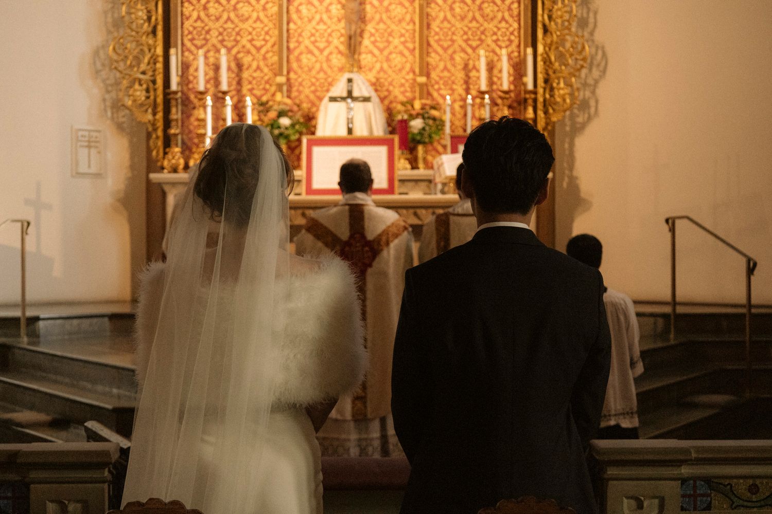 Bride and groom stand before ornate golden altar with candles during traditional Catholic wedding Mass.