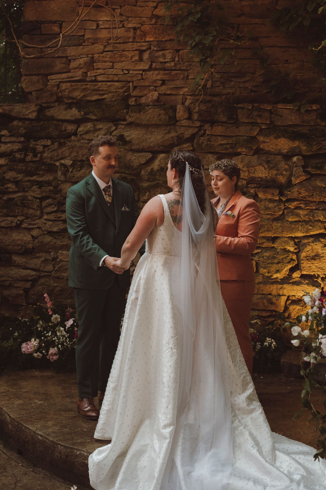 A wedding ceremony takes place against a rustic stone wall backdrop with floral arrangements and a flowing white bridal gown.