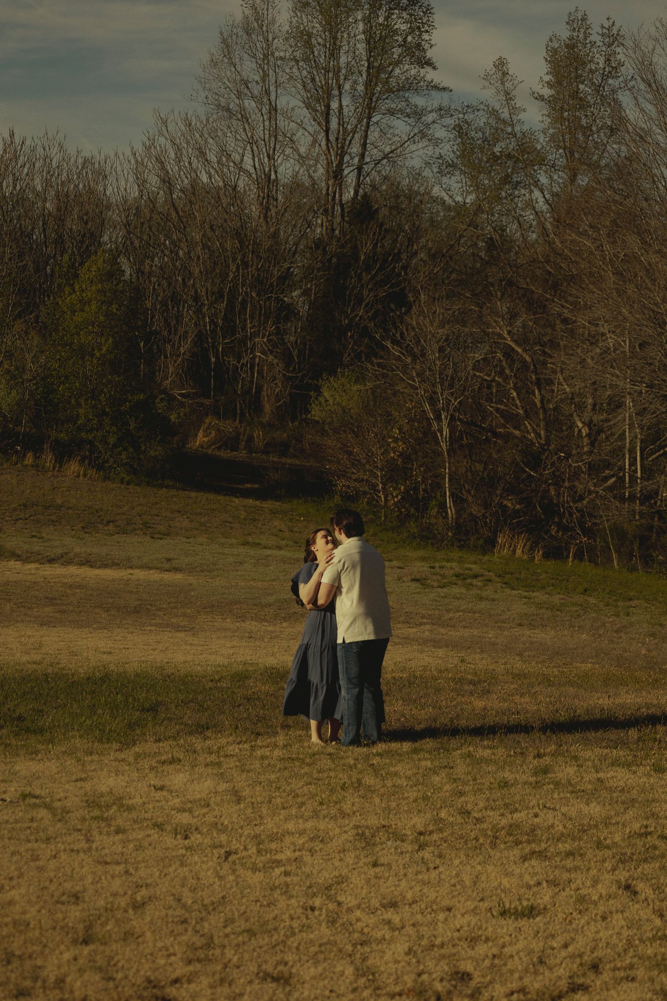 Couple embracing in a golden sunlit field surrounded by bare autumn trees.