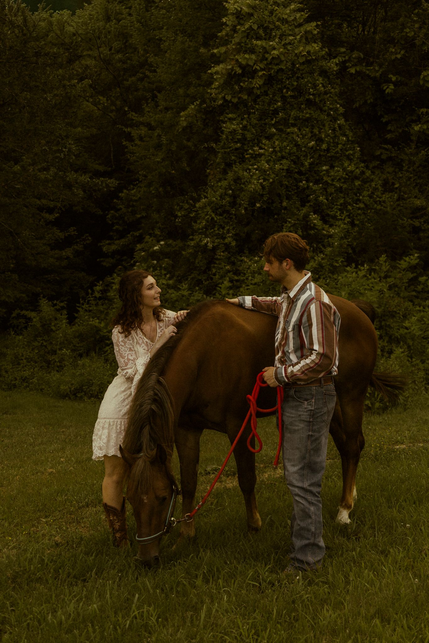 A couple stands with a brown horse in a lush green field, woman in white dress and man in striped shirt holding red lead.