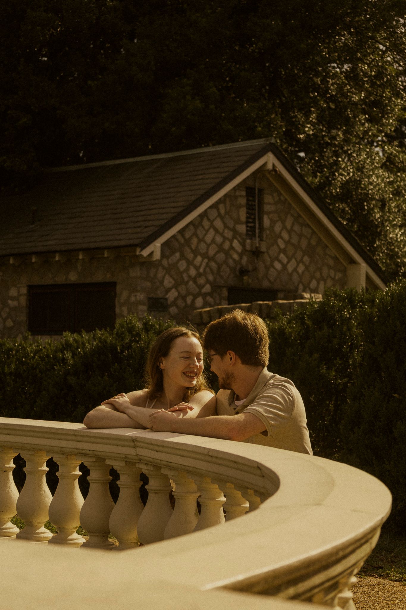 A young couple shares a romantic moment leaning on a white balustrade railing outdoors near a stone building.