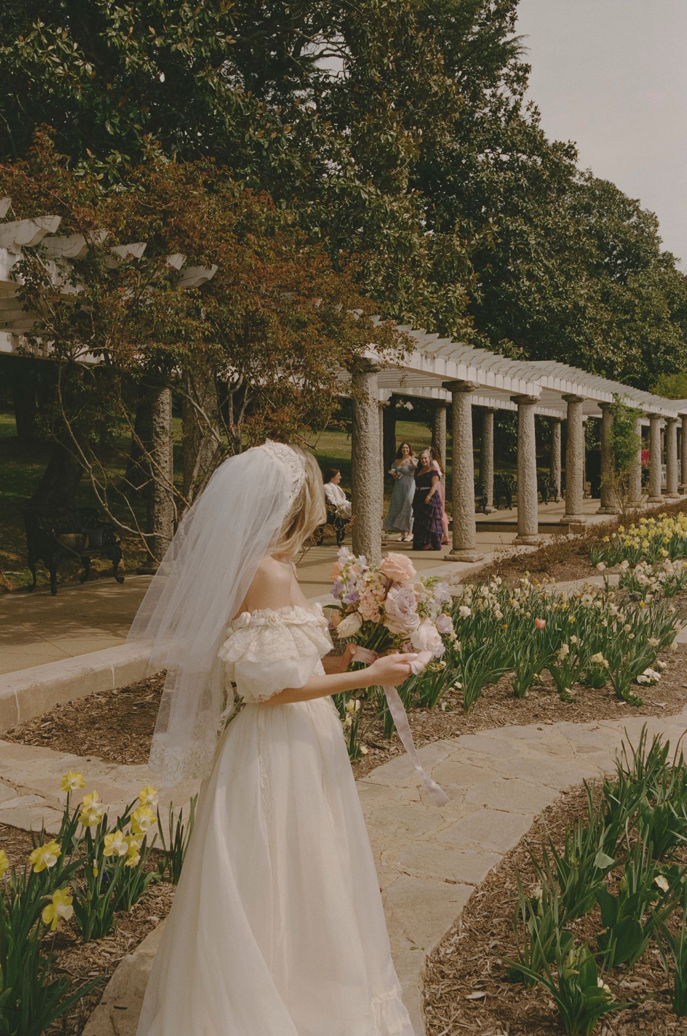 A person in a flowing white dress walks along a garden path bordered by columns and spring flowers in bloom.