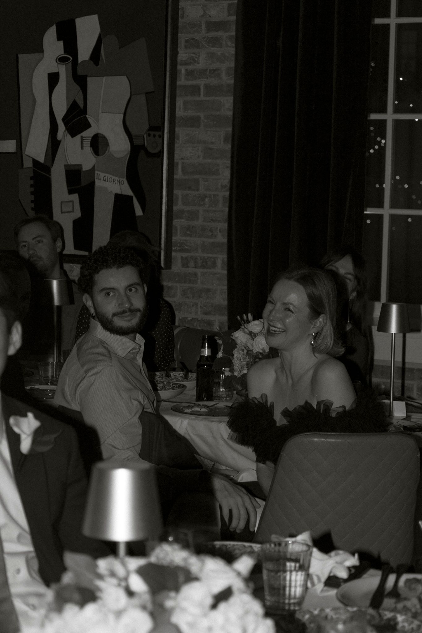 Black and white photo of smiling wedding guests seated at a reception dinner table with floral centerpieces.