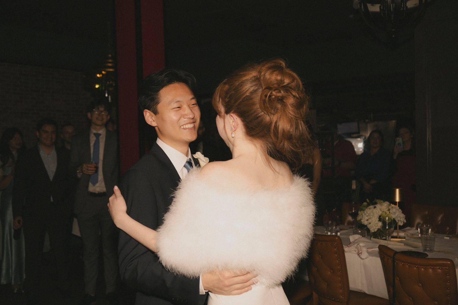 Groom in suit smiles while dancing with bride in white fur wrap at wedding.