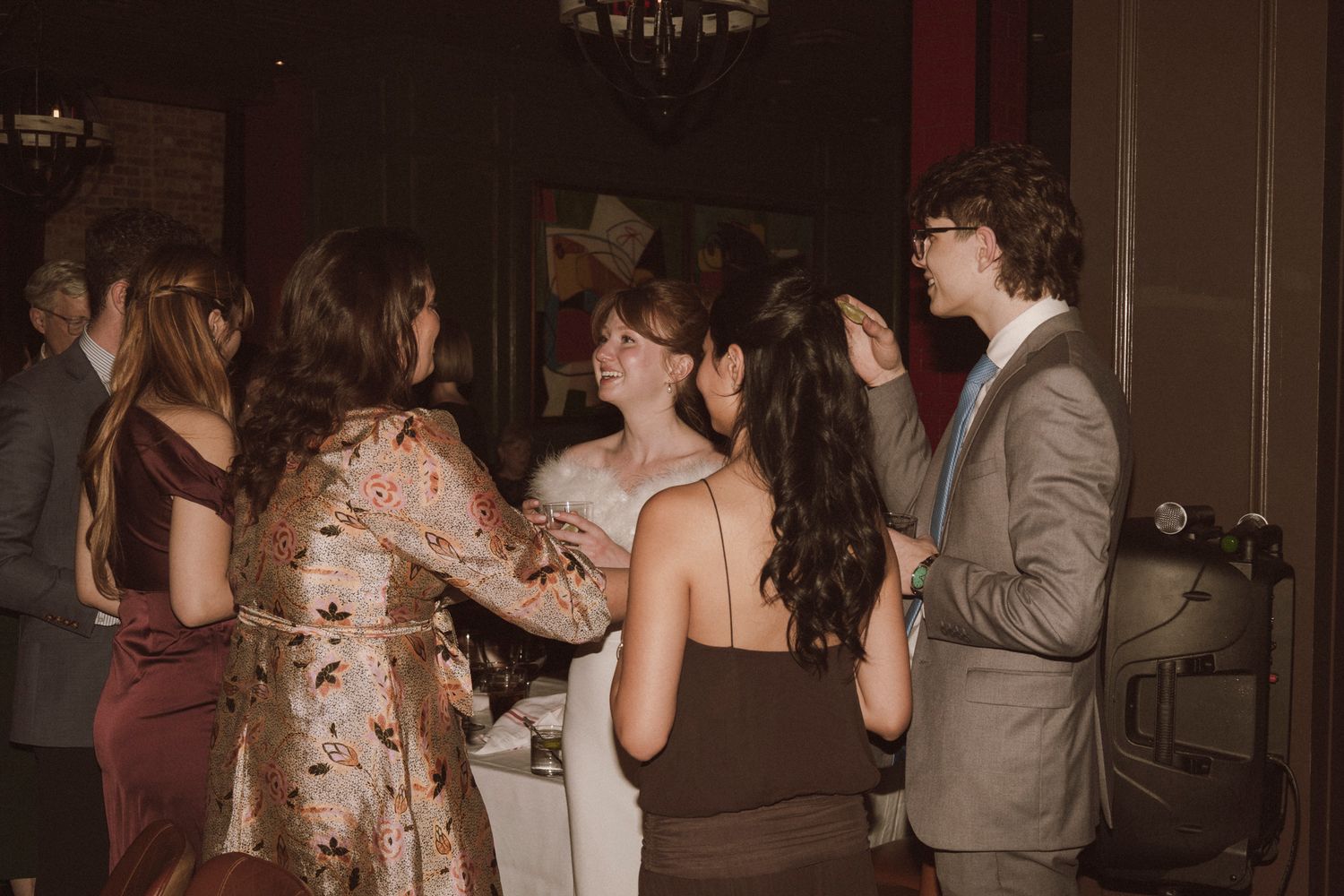 Group of young adults socializing and dancing at a formal event, wearing elegant attire in warm tones.