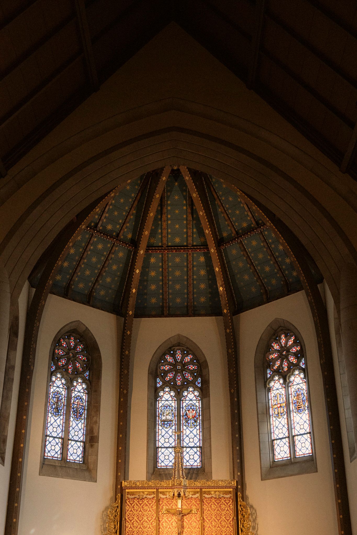 Church apse with octagonal ceiling, colorful stained glass windows, and golden altar cross.