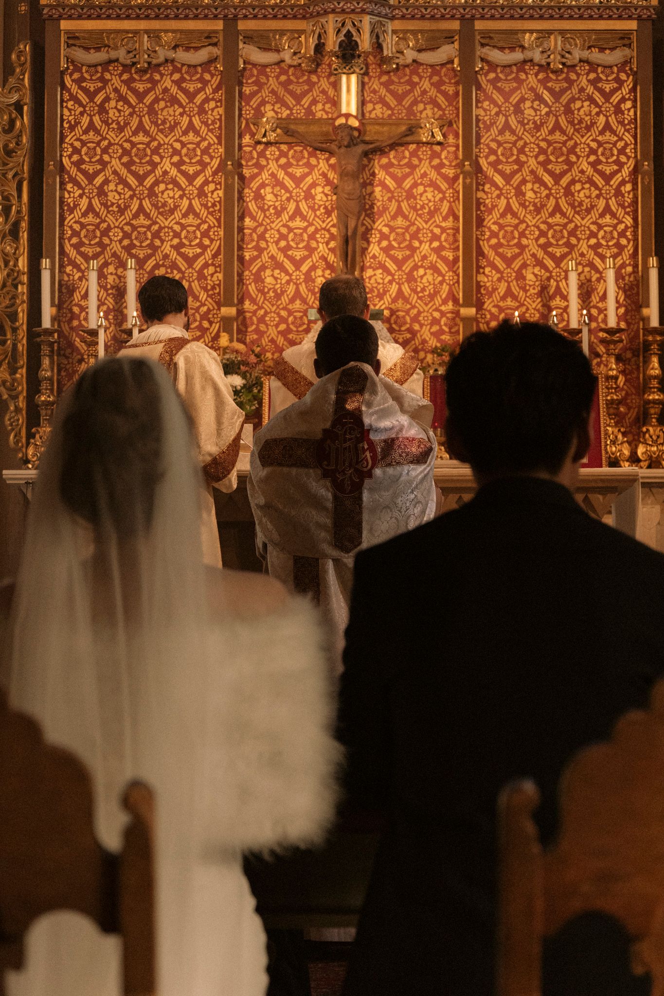 Bride and groom kneeling before priest at ornate golden altar during Catholic wedding.