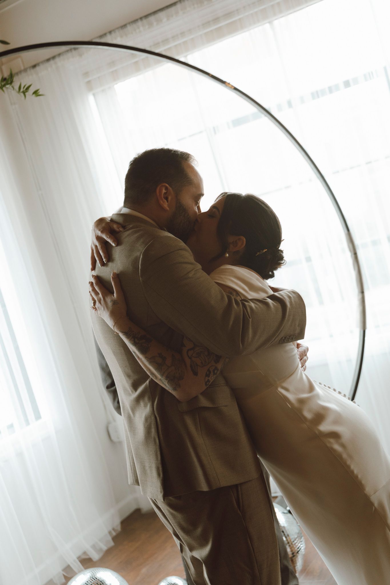 A romantic wedding couple embraces near a sunlit window with soft white curtains.