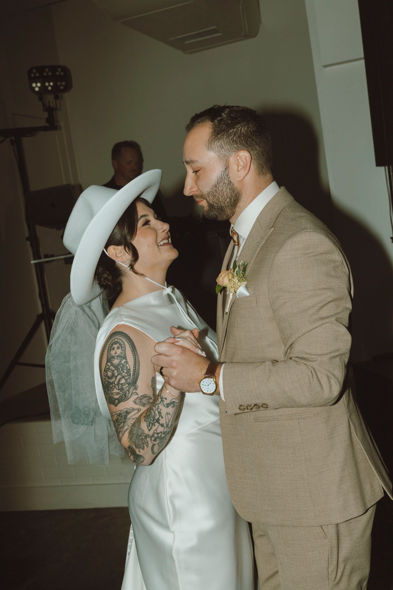 A couple shares their first dance at their wedding, with the bride wearing a white hat and dress showing tattoo sleeves.