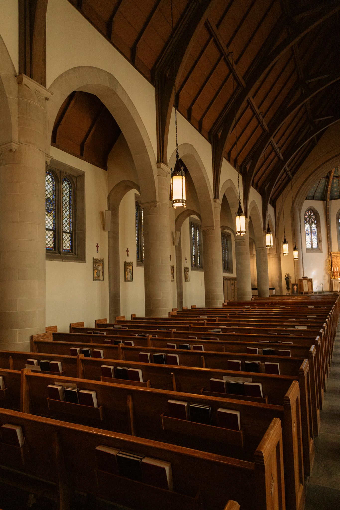 Gothic church interior with wooden pews, stone arches, and hanging lanterns along the nave aisle.