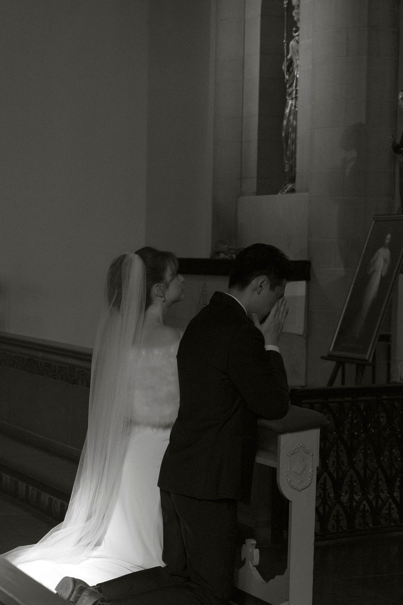 Black and white photo of bride and groom kneeling in prayer at church altar during wedding ceremony.