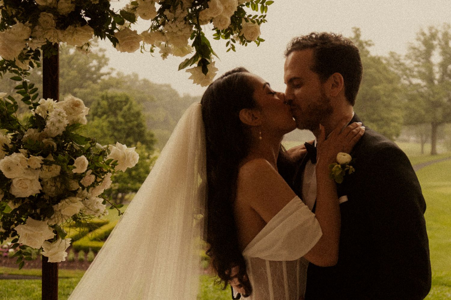 Bride and groom share a romantic first kiss under a floral arch with white roses during an outdoor wedding ceremony.
