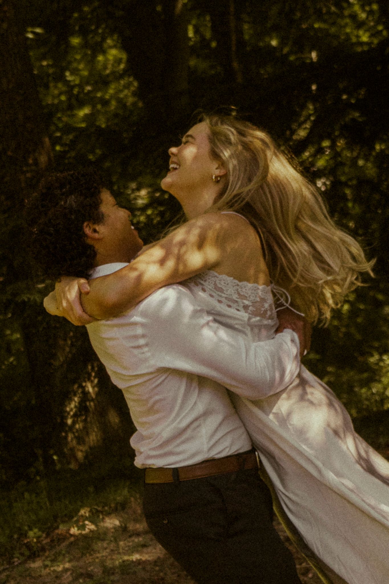 A joyful bride in a white gown laughs as her groom lifts her in a romantic forest wedding photo.
