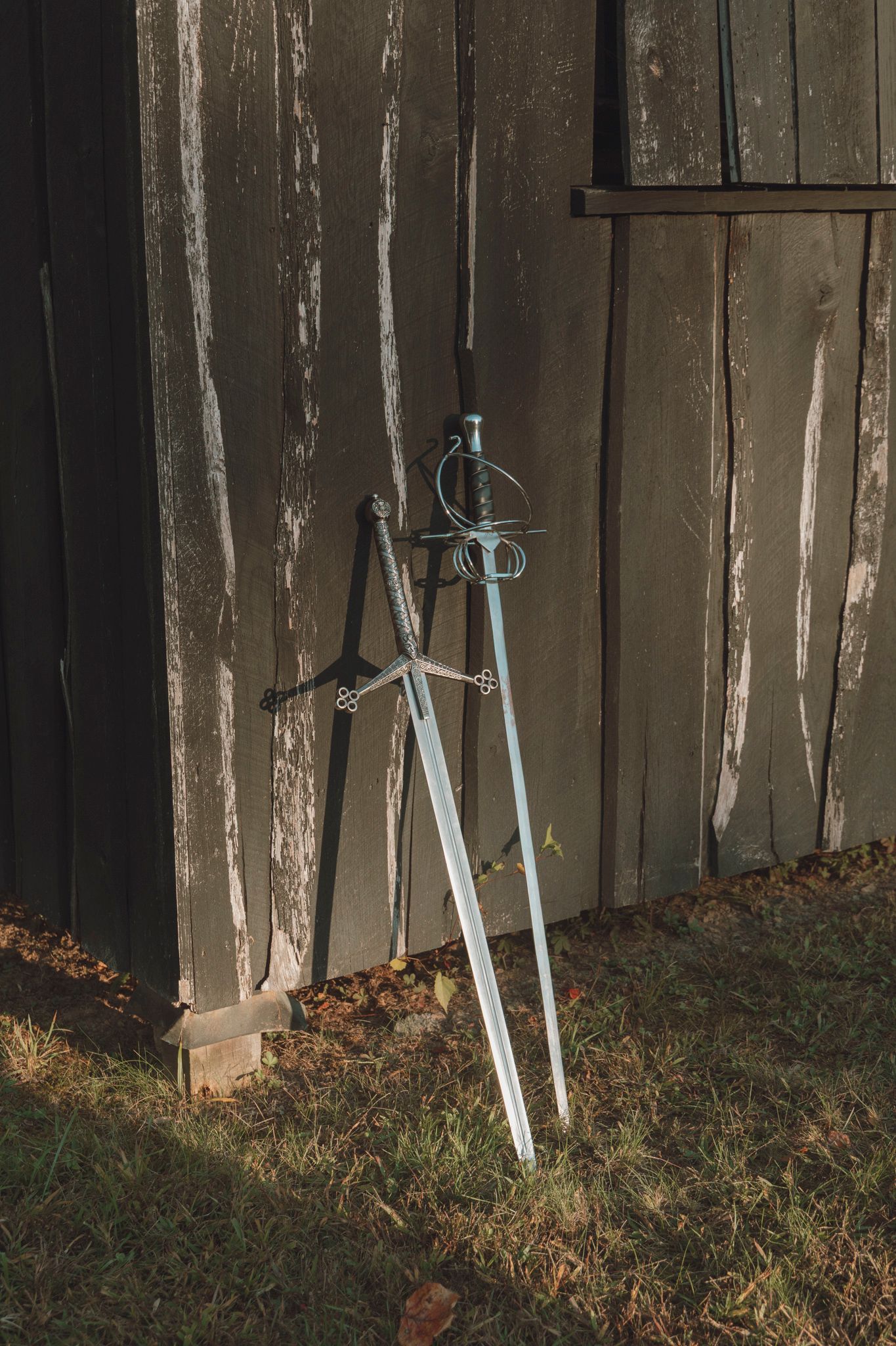 Three decorative swords lean against a weathered wooden fence in a garden setting with fallen leaves.