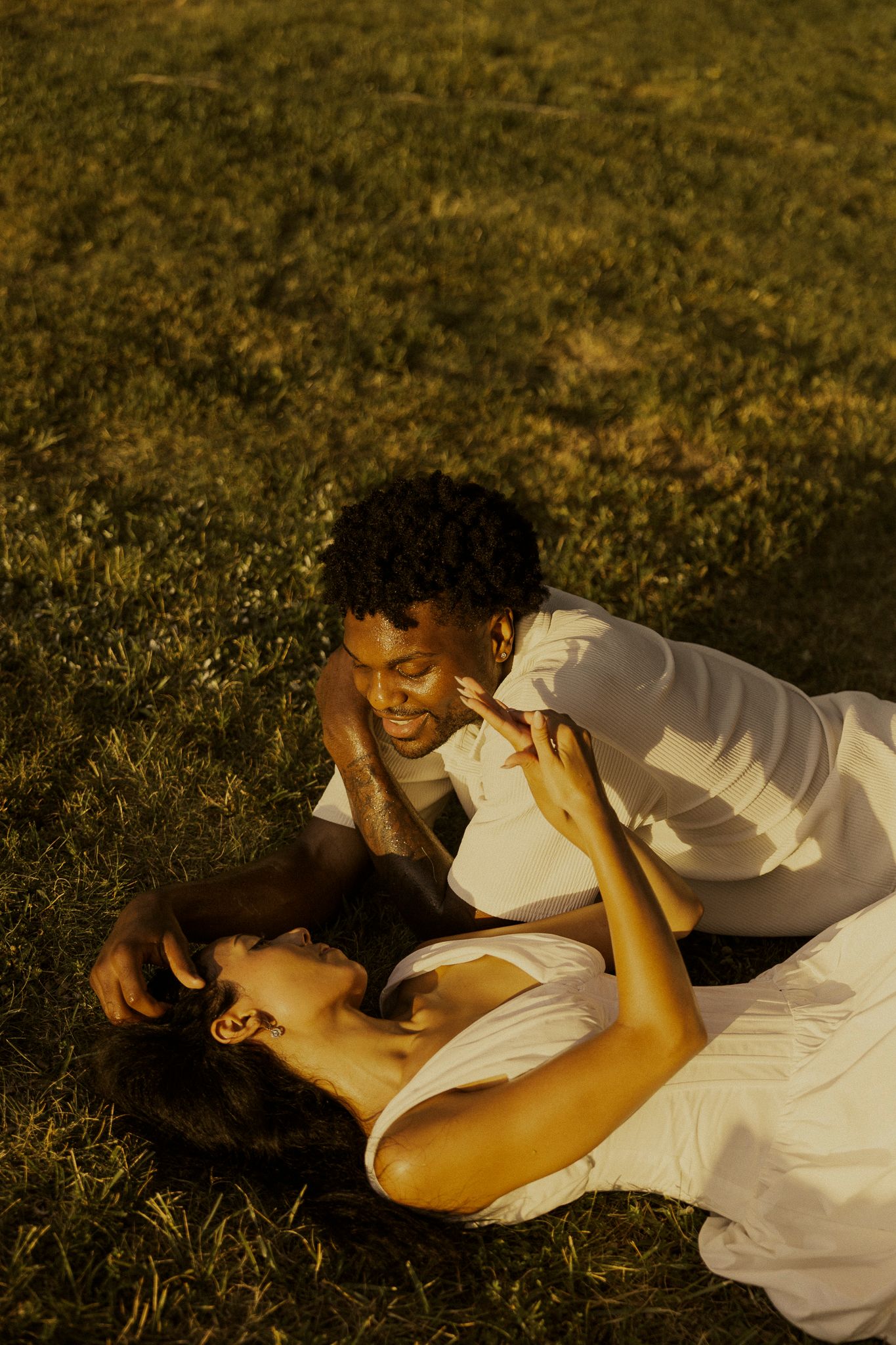 Couple sharing a romantic moment on green grass, woman in white dress lying down as man smiles at her warmly.