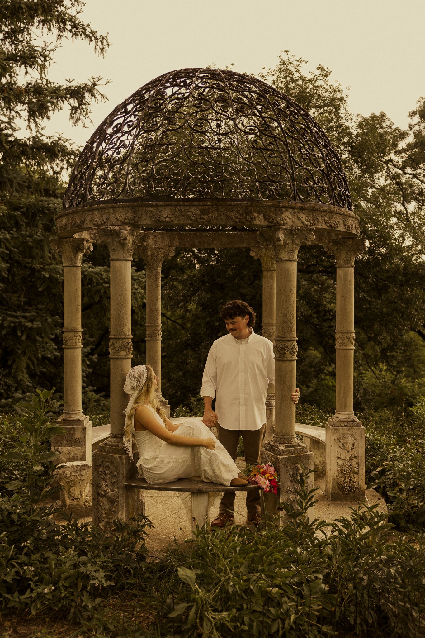 Bride and groom share a romantic moment inside an ornate stone gazebo surrounded by lush greenery.