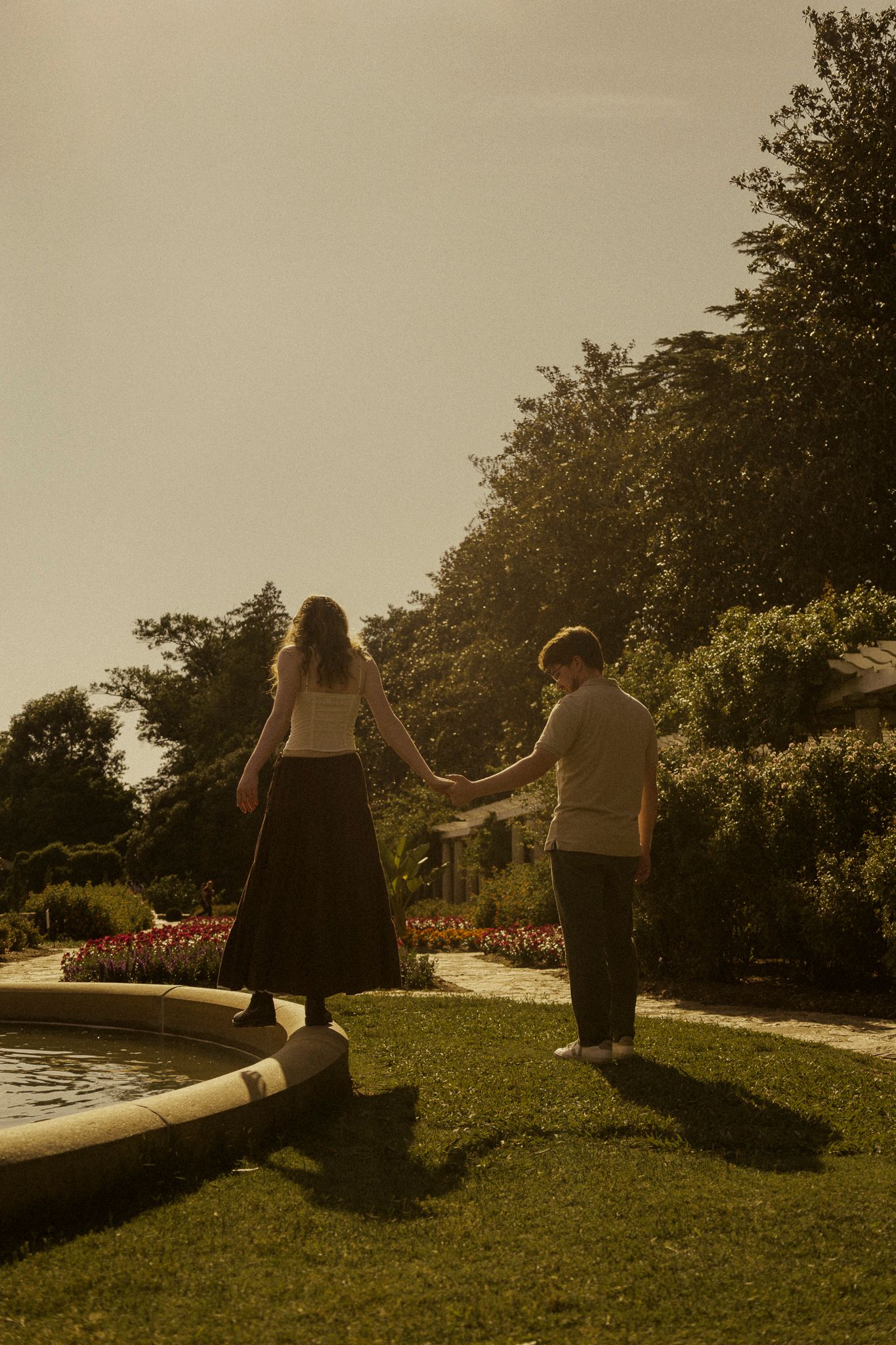 Couple holding hands walking by a garden fountain surrounded by lush greenery and colorful flowers in warm golden light.