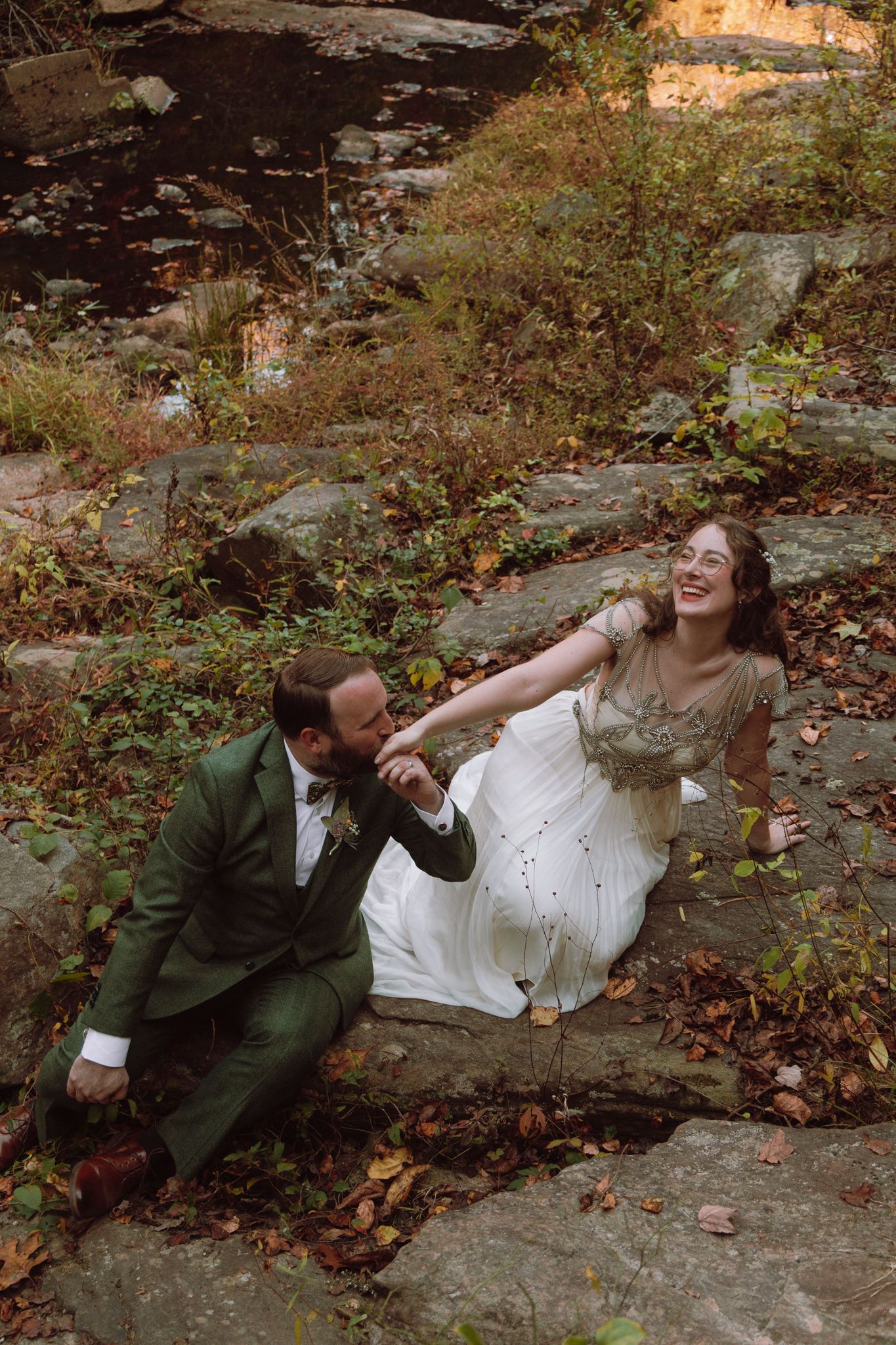 A couple in wedding attire share joyful moments while laying on rocks surrounded by autumn leaves.
