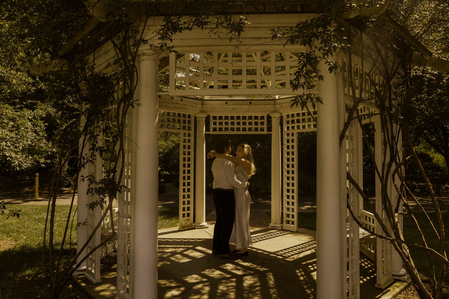 Couple embracing under a white lattice garden gazebo surrounded by lush greenery in warm golden light.