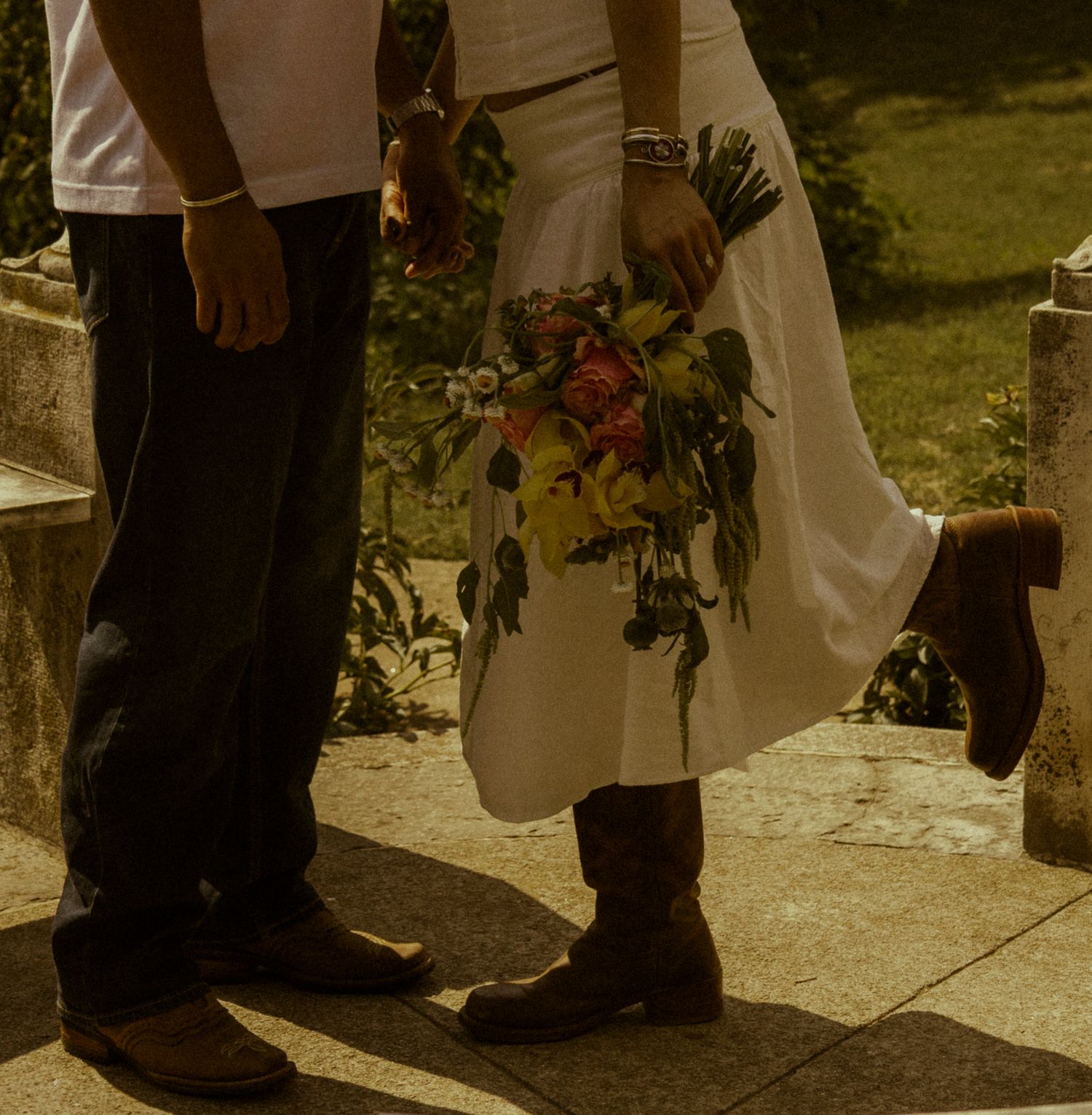 Bride in white dress and cowboy boots holding colorful bouquet standing beside groom in dark pants and brown shoes.