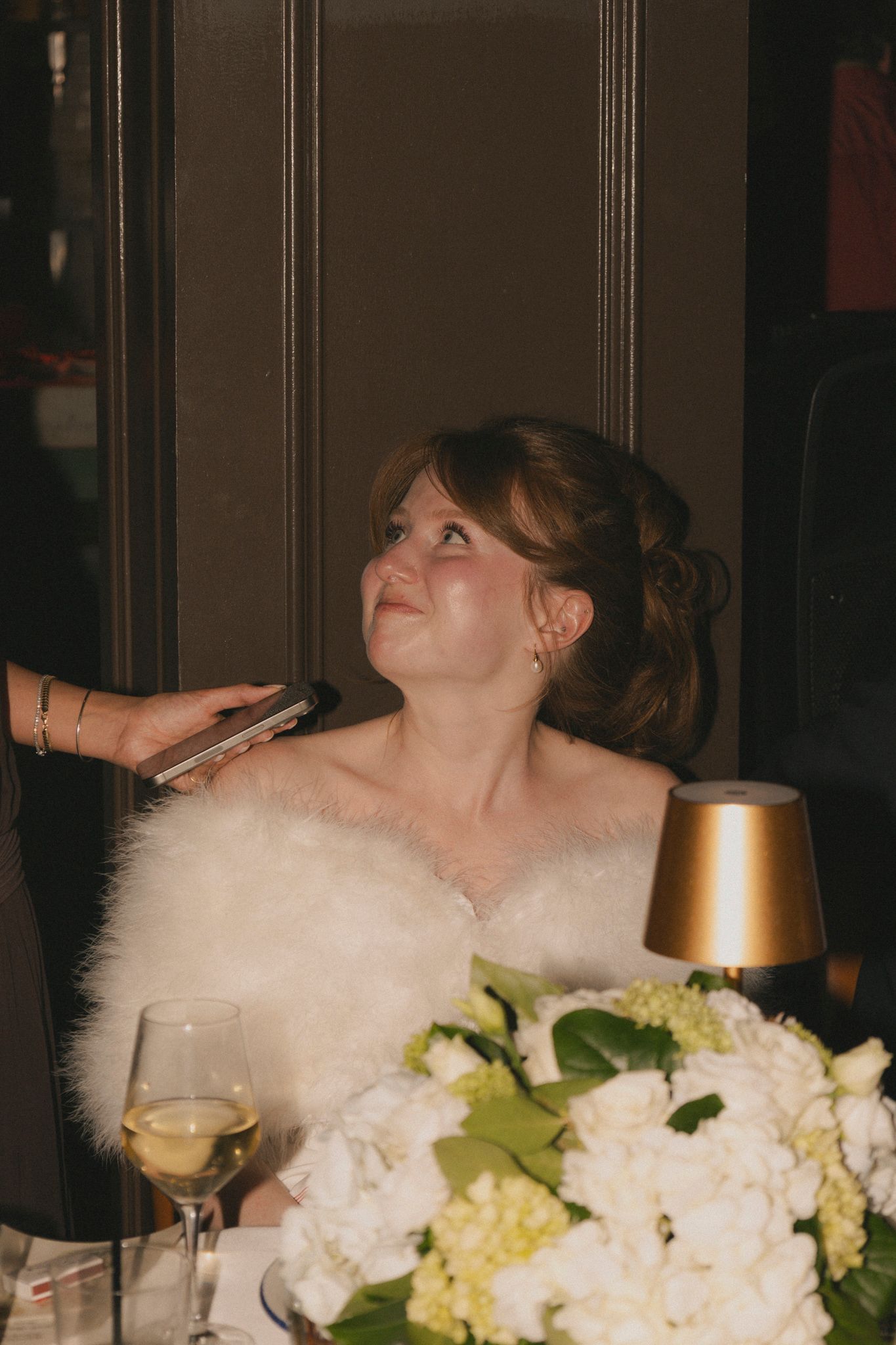 Emotional bride in white fur stole looks up tearfully during wedding reception speeches, with floral centerpiece nearby.