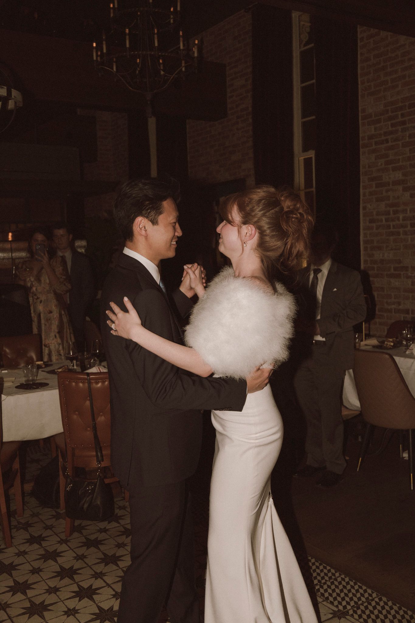 Bride and groom sharing first dance in elegant brick-walled reception venue with chandelier.